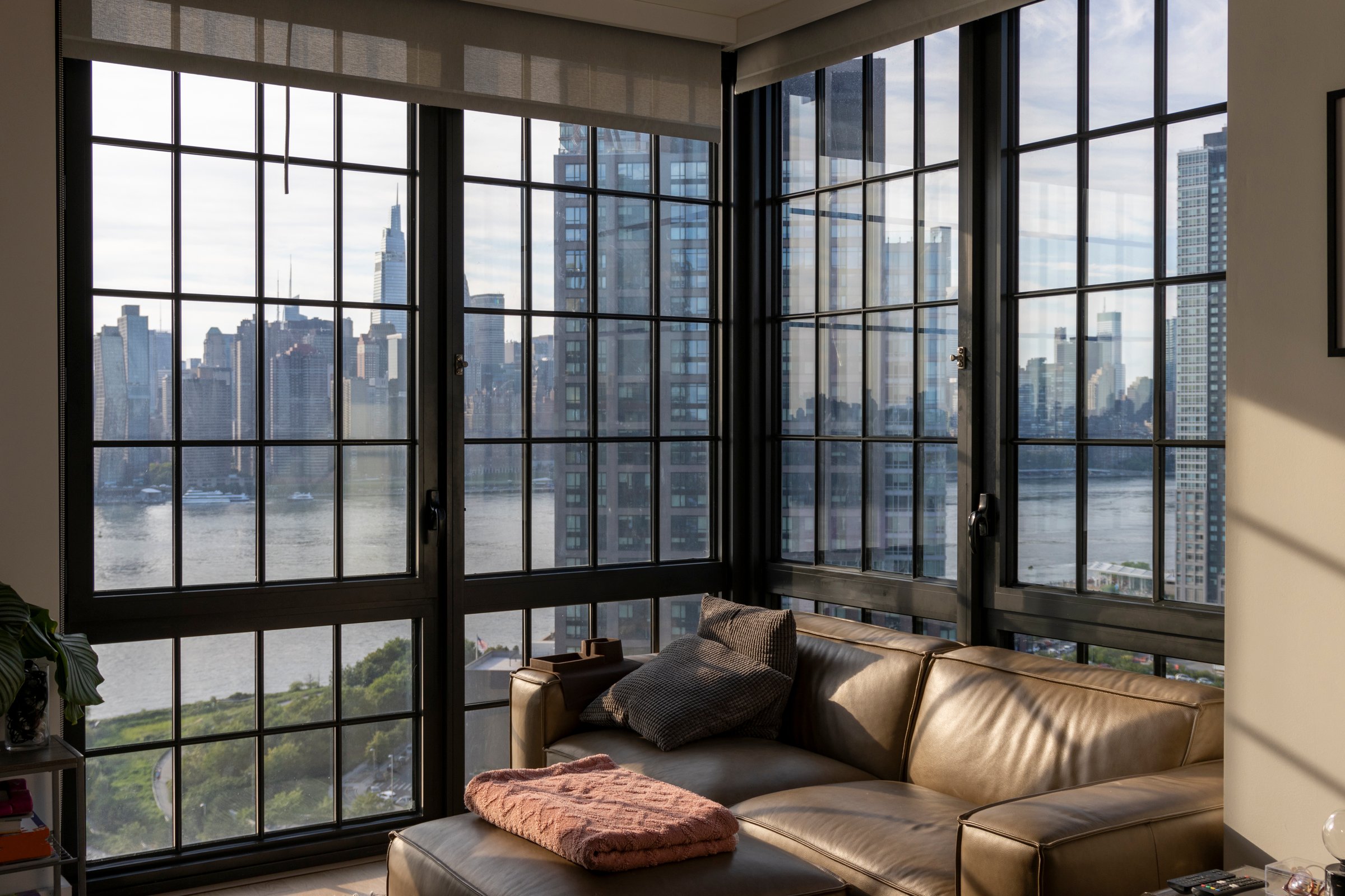 The spacious living area features modern furniture and large windows that frame a stunning view of the New York skyline, bathed in soft afternoon light.