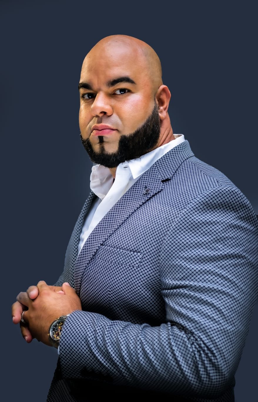 Man in a gray checkered suit and white shirt posing against a dark background, with hands clasped and a watch visible.