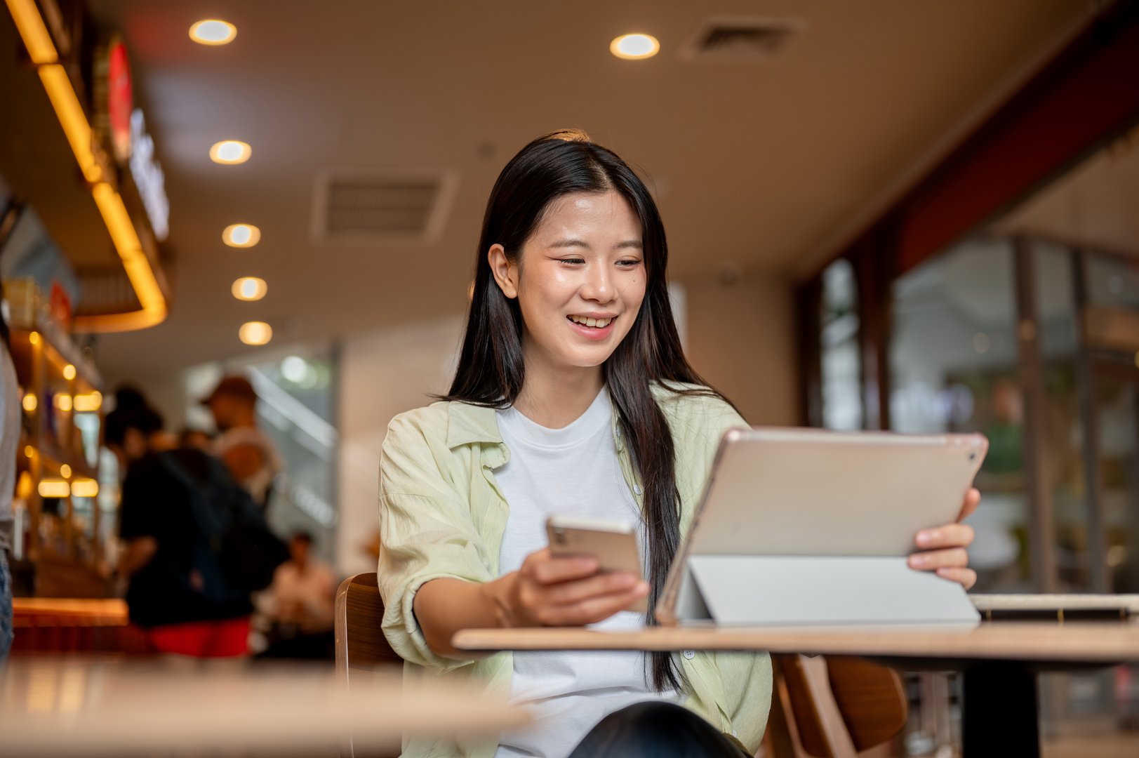 Asian woman student laughing while looking at tablet and holding phone on wooden table in the cafe. Online Entertainment, Modern tech, Urban City Life