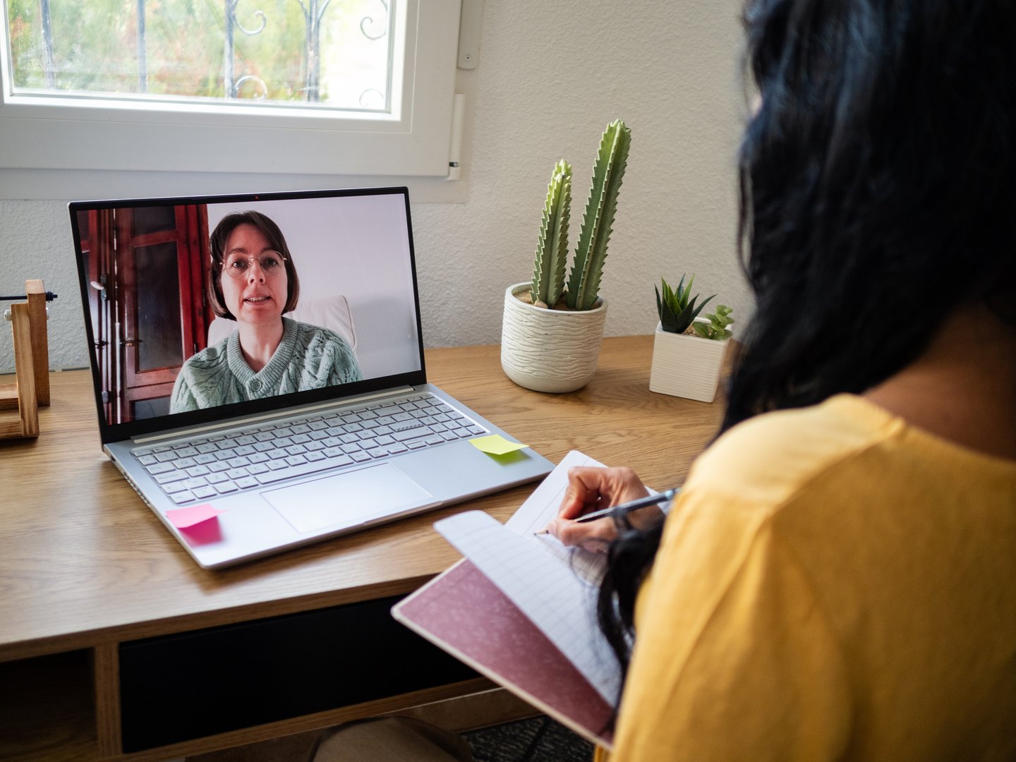 Video call session between a patient and a Mexican female psychologist. She appears focused as she takes notes, surrounded by a simple, calm workspace with plants.