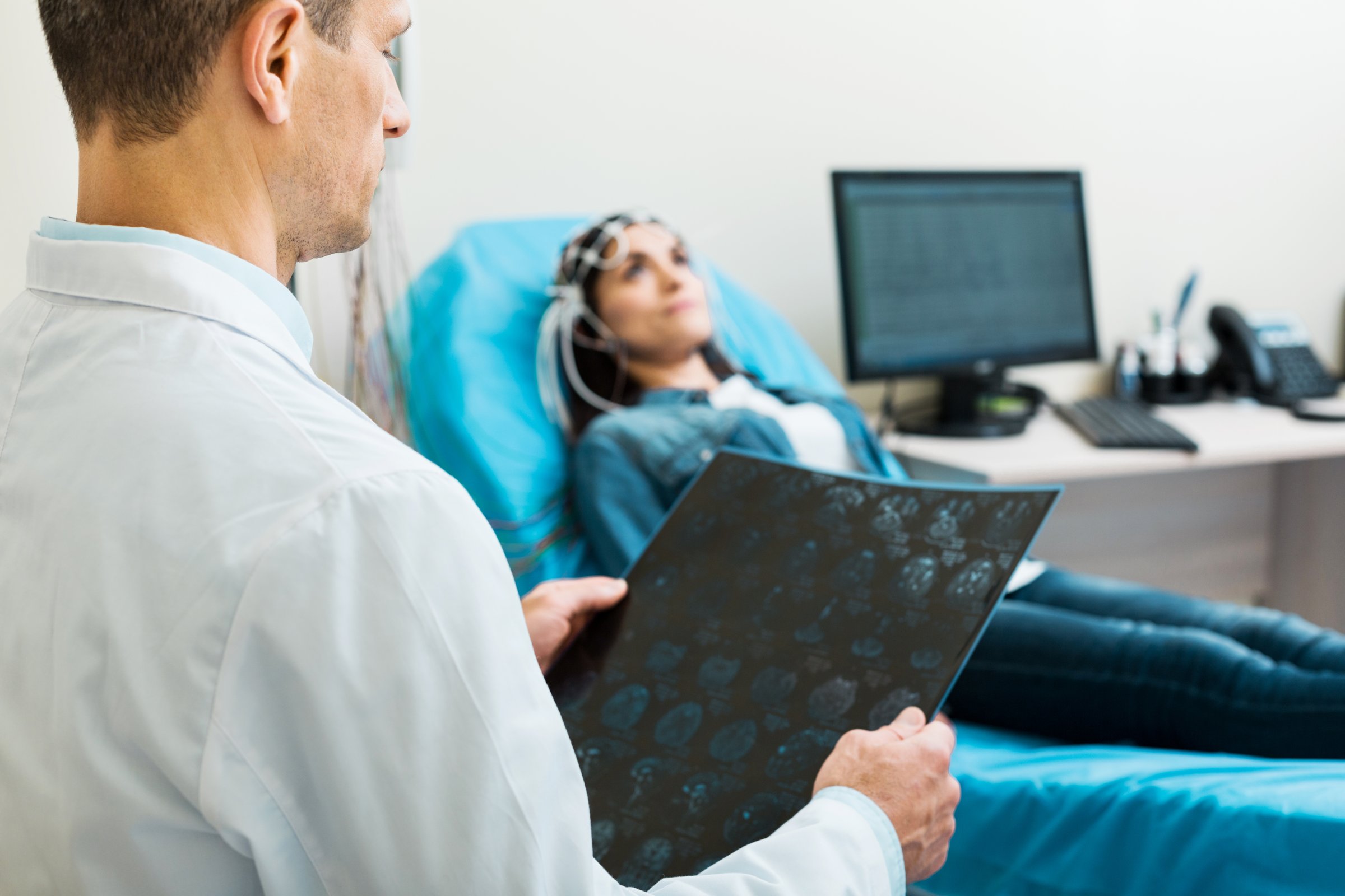Considering all details. Pleasant middle-aged man examining the results of computer tomography and carrying out electroencephalography of a young woman in the meantime