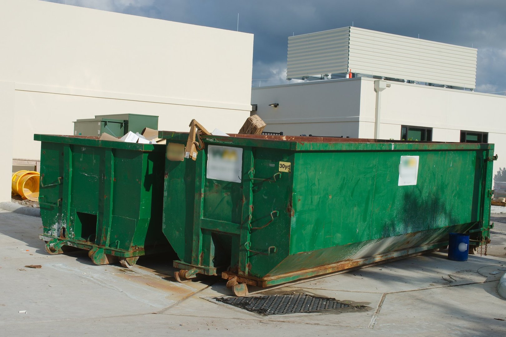 Photograph featuring two large green industrial dumpsters situated near a white building exterior under a cloudy sky. The urban setting suggests waste management or construction cleanup activities.