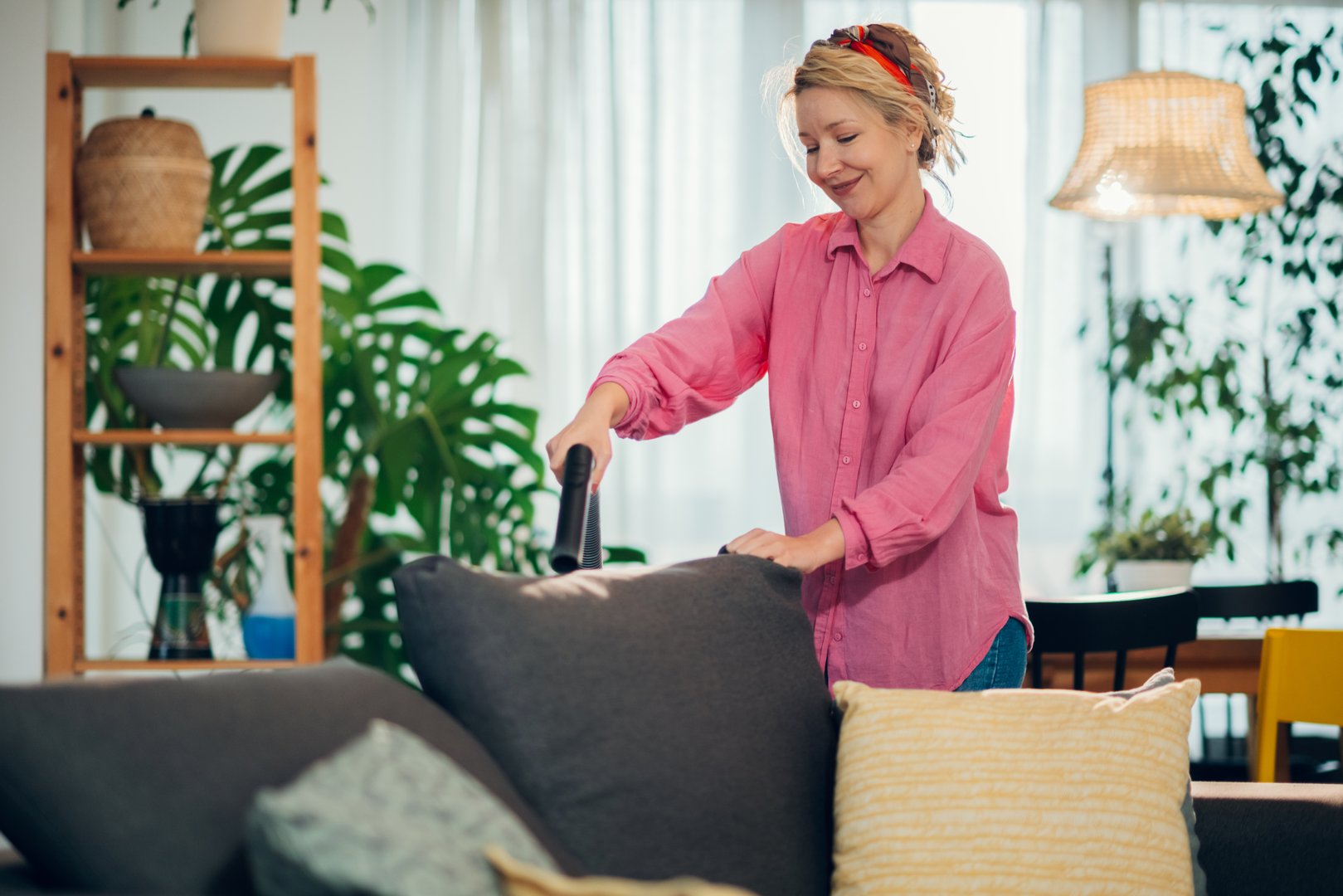 Smiling housewife using handheld vacuum cleaner to remove dust and mites from sofa in living room, enjoying domestic chores and keeping her home clean and tidy