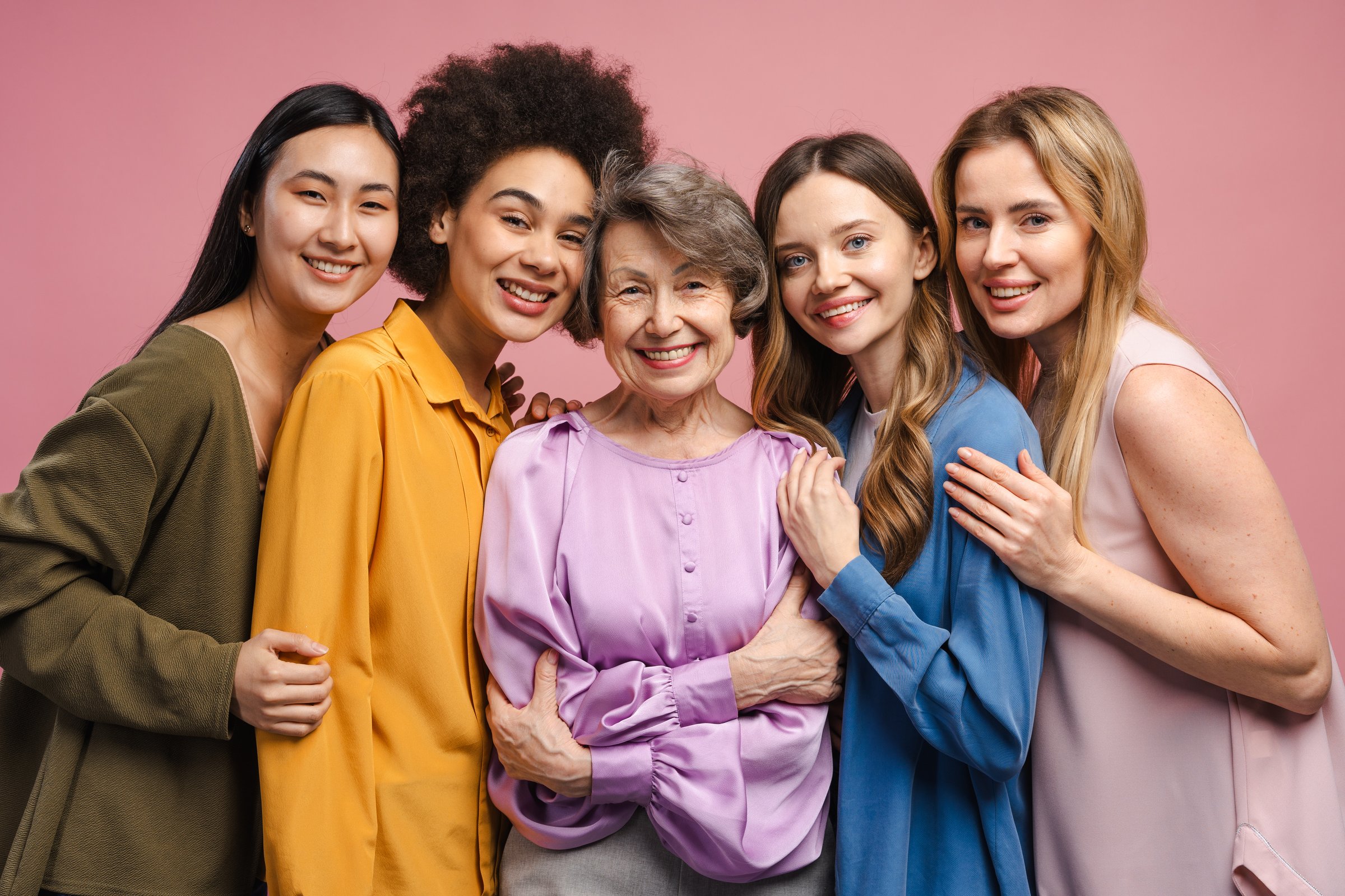 Five cheerful multiethnic women of different ages and jobs are hugging each other and smiling on a pink background, showing unity and female empowerment