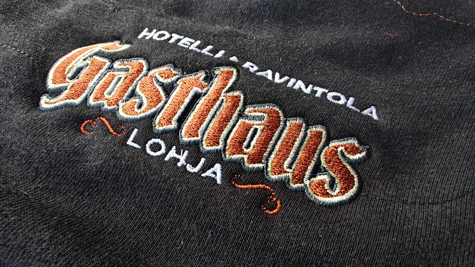 Embroidered text on fabric reading Hotelli Ravintola Gasthaus Lohja with decorative orange stitching.