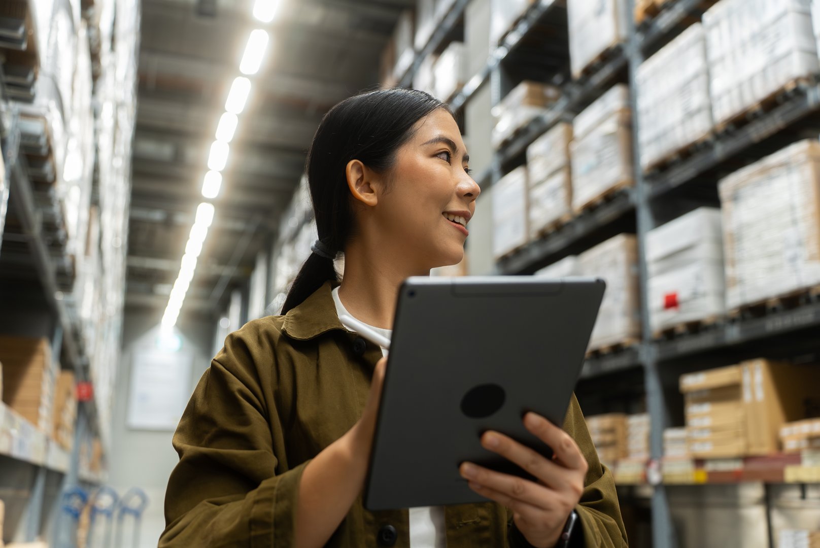Smiling woman worker look around the stacked shipping boxes while holding smart digital tablet to track real-time stock inventory in logistics center and supply chain products in warehouse.