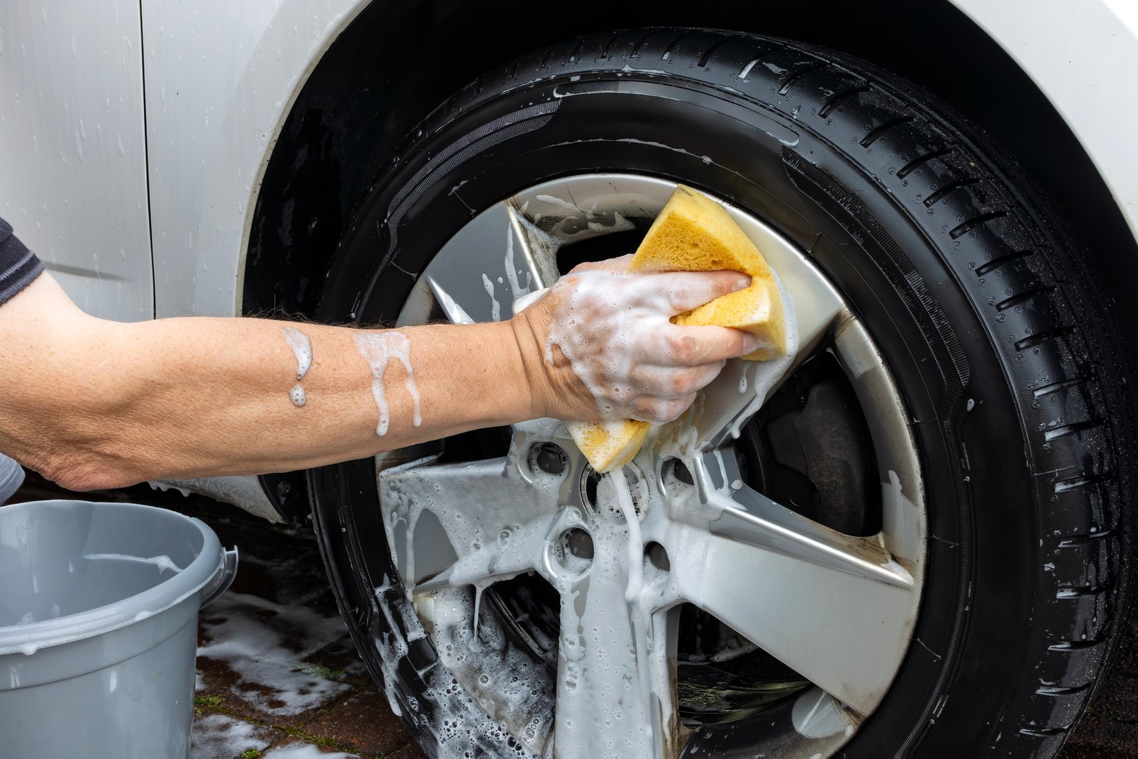 Close-up of a person's hand washing a silver alloy wheel with a yellow sponge and soapy water.