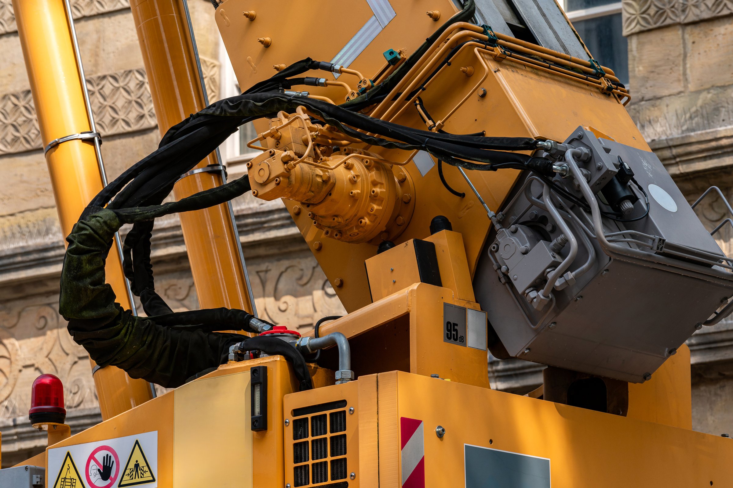 A robust machinery arm is lifted high as it engages in construction activities in an urban setting. The equipment features various warning signs and is positioned near a historical building.