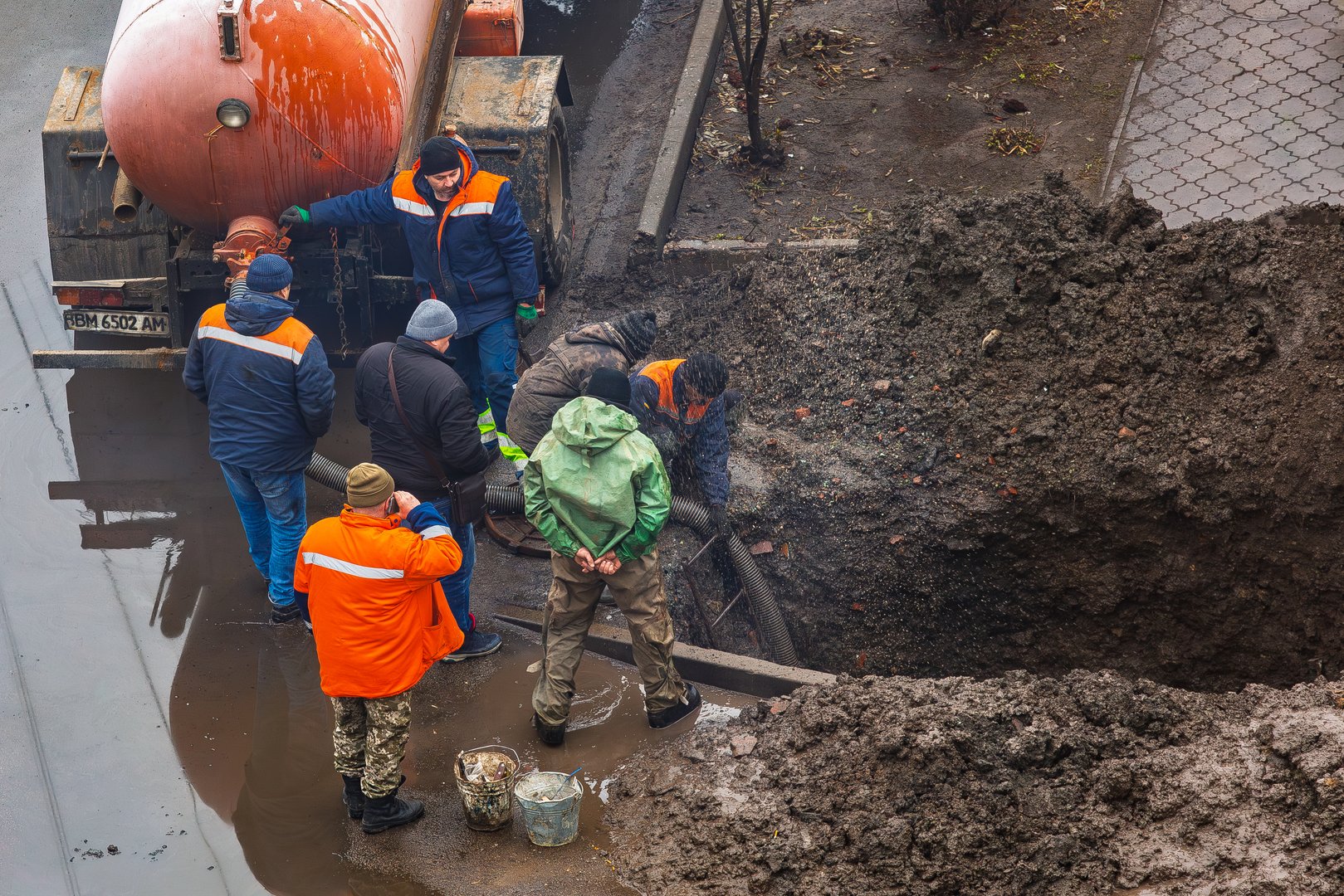 Ukraine, Romny, December 24, 2024: A group of workers repair a damaged pipeline in a muddy trench with a sewage truck parked nearby. The workers are surrounded by tools, water and mud.