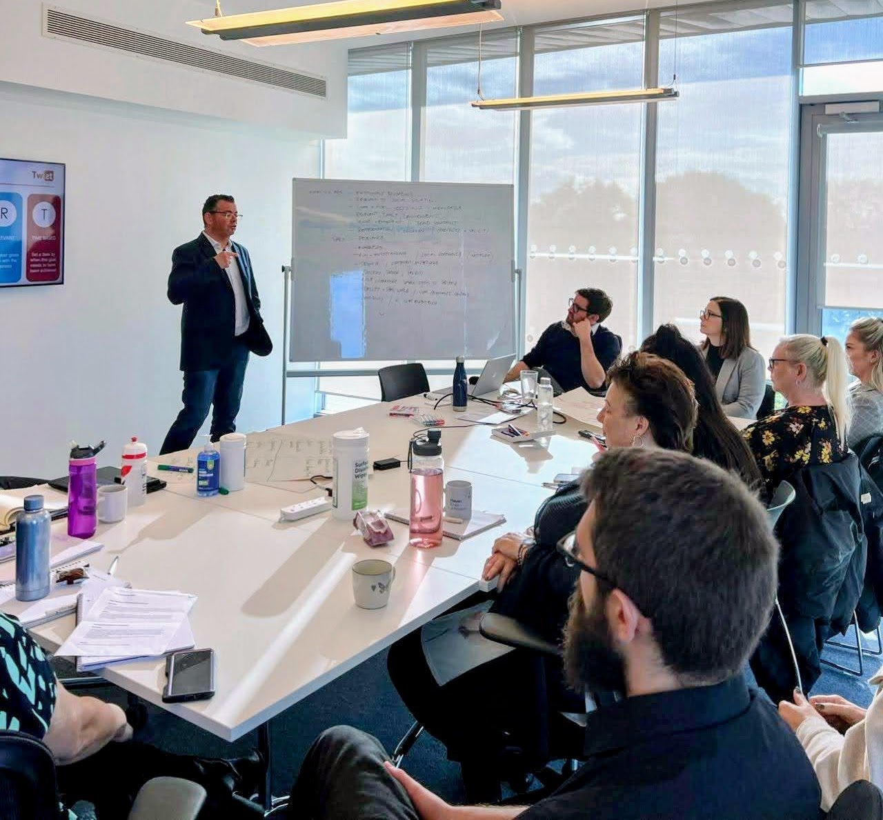 A man presents at a whiteboard to a group seated around a conference table. Notes and drinks are on the table.