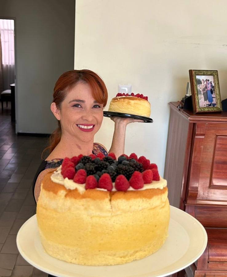Woman smiling, holding a small cake with berries, with a larger similar cake in front of her on a table.