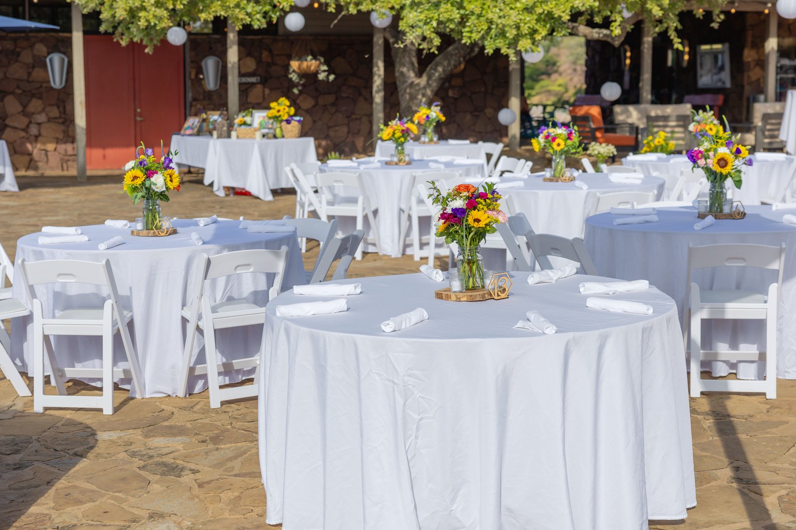 Outdoor wedding reception setup with round tables covered in white tablecloths, adorned with colorful floral centerpieces, under a sunny sky.