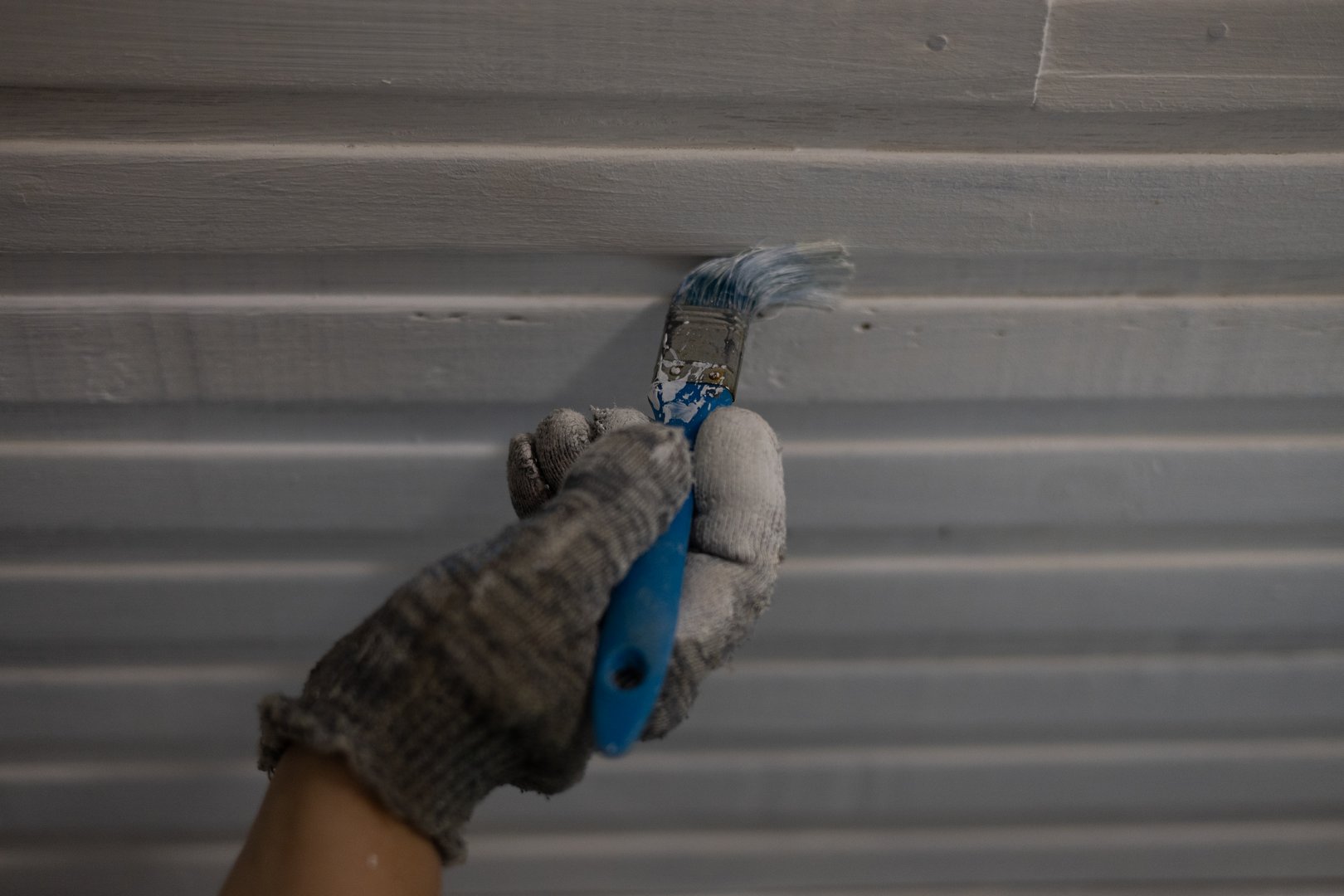 Close-up of a painter's hand wearing gloves applying white paint on a wooden ceiling using a brush, demonstrating a precise and professional painting technique during a home renovation project..