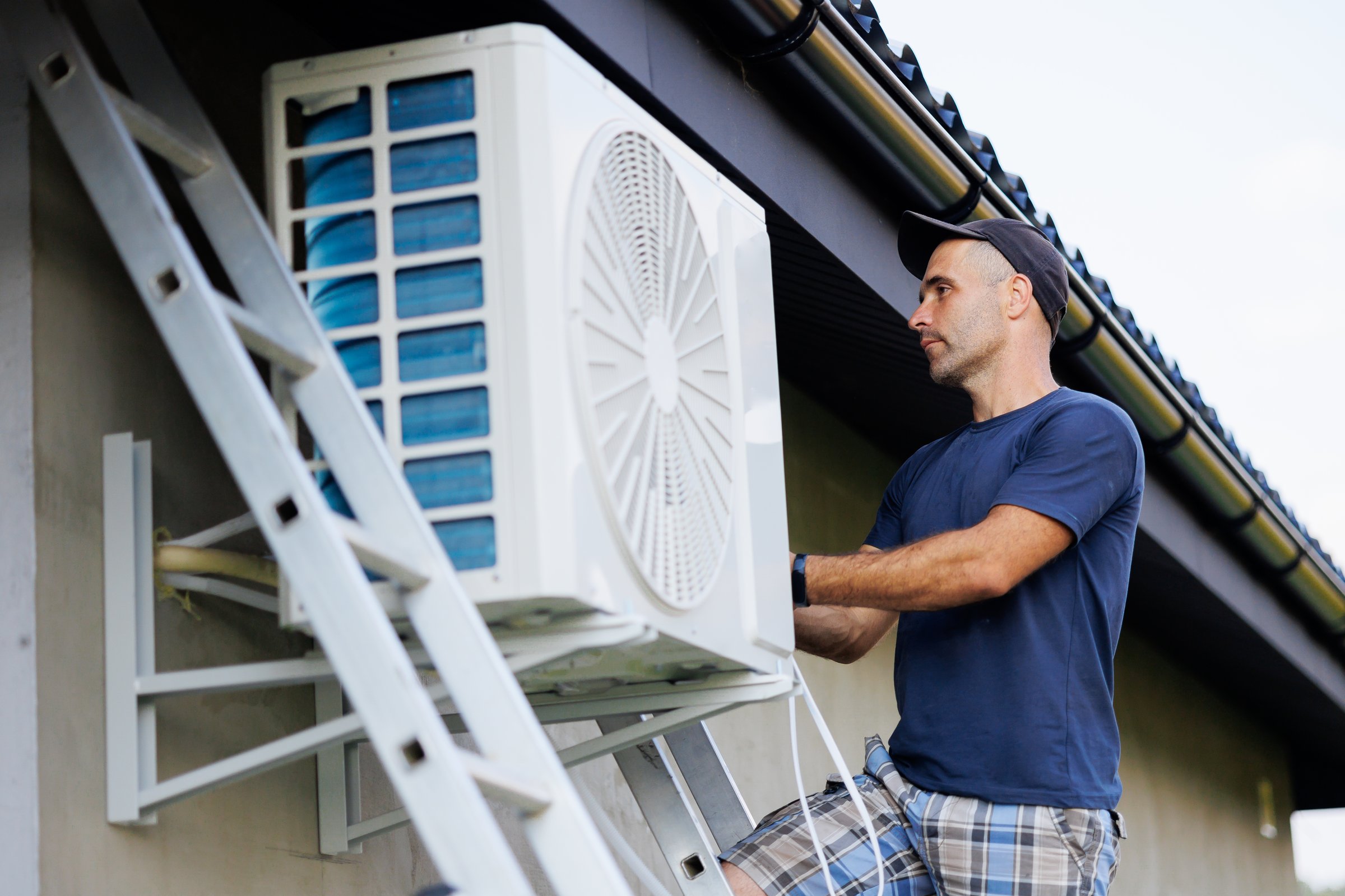 A man is working on an air conditioner. He is wearing a blue shirt and plaid pants