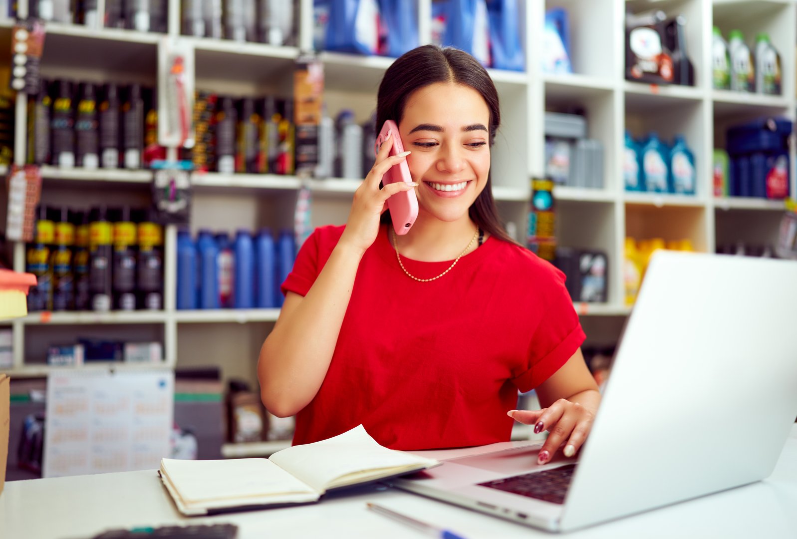 Smiling saleswoman managing orders by phone and entering data into laptop in auto parts store