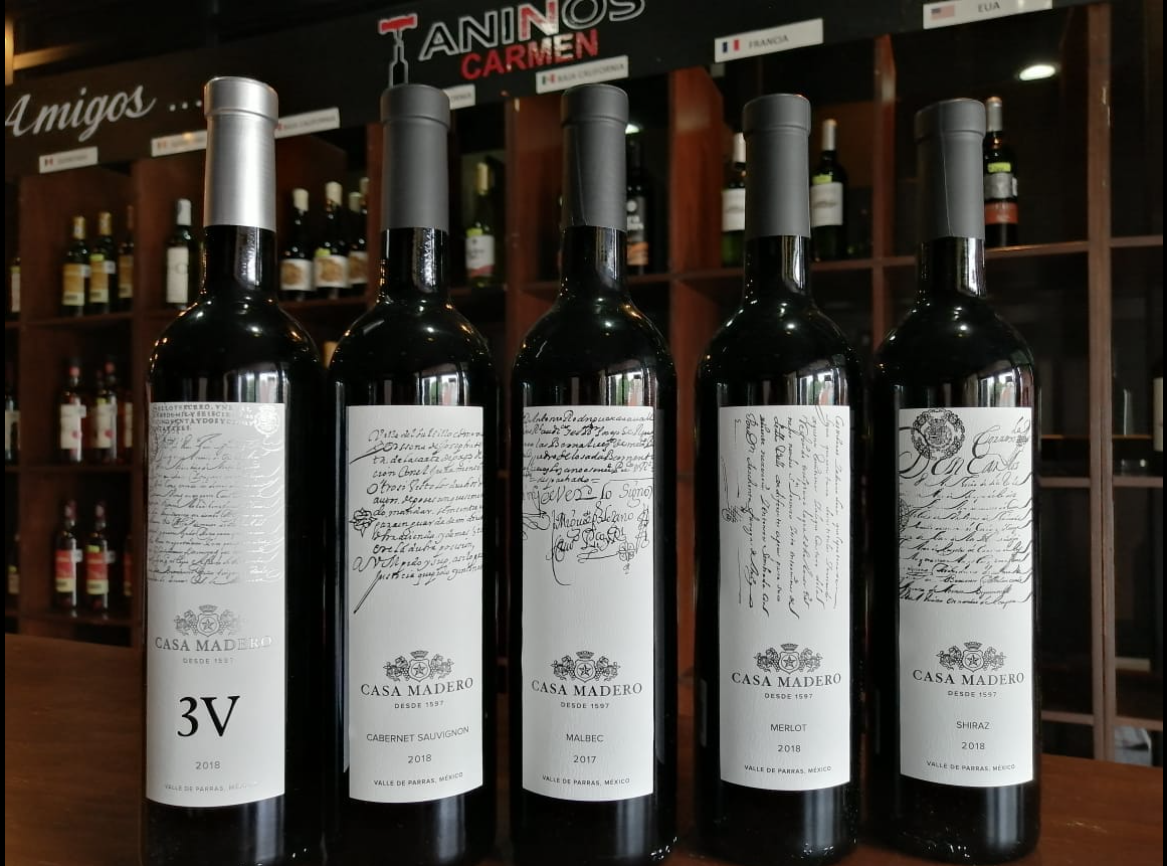 Five bottles of Casa Madero red wine on a wooden shelf with handwritten labels displayed in a wine shop.
