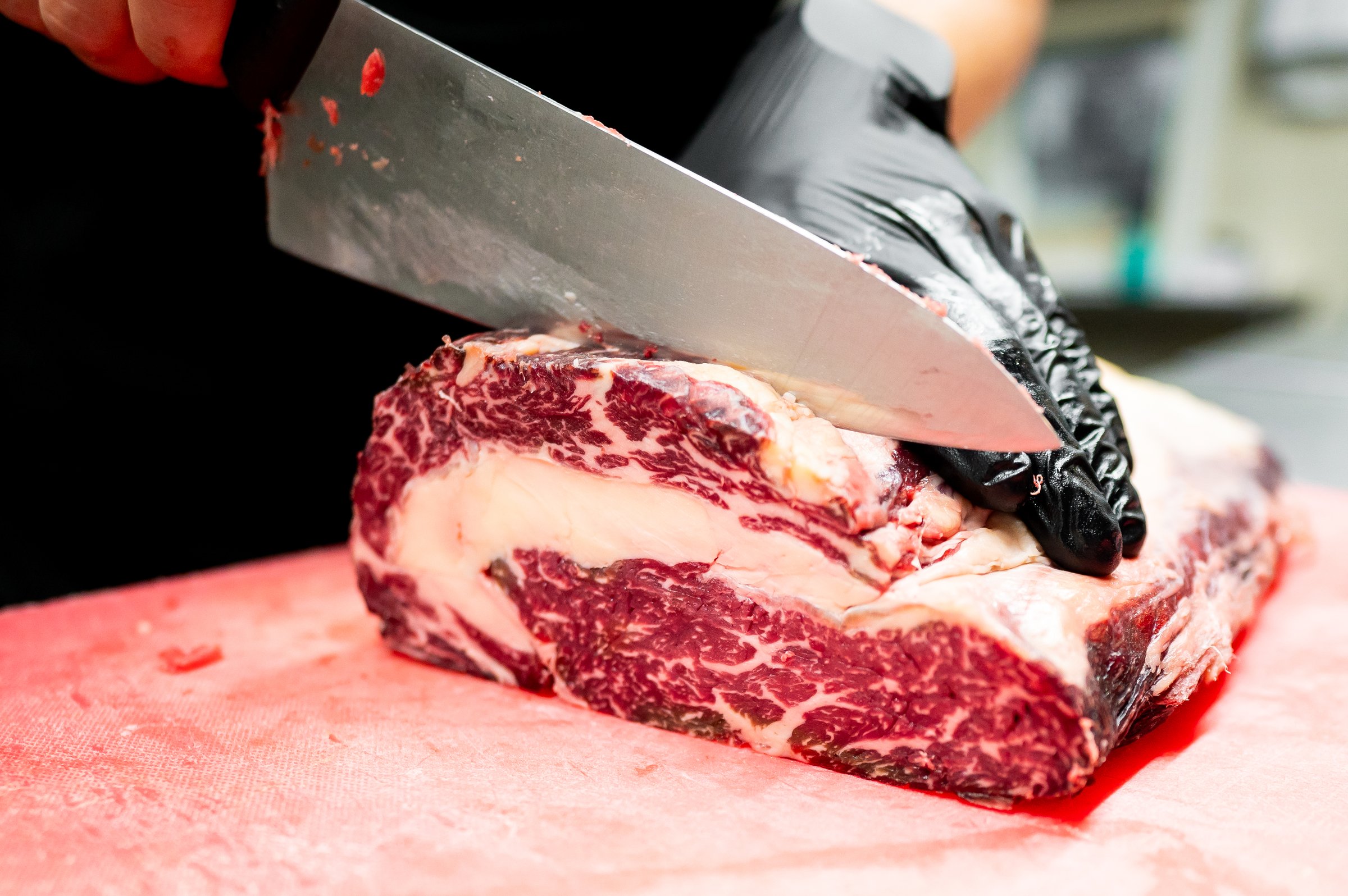 close-up of a hand slicing raw steak on a red cutting board with a sharp knife. image focuses on the precise action of cutting, highlighting the texture of the meat and the cleanliness of workspace.