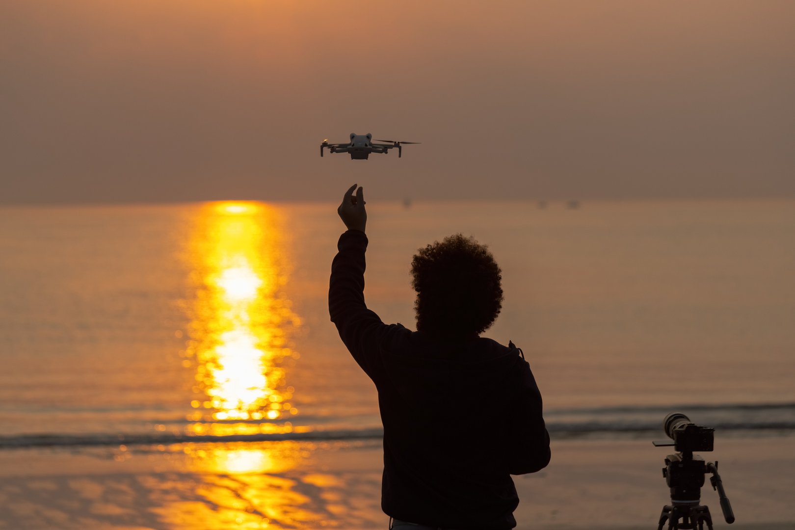 Man backview unrecognized launching drone with a camera on tripod next to him on the beach at sunrise