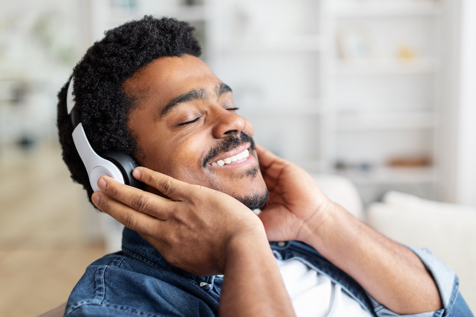 African American man with a joyful expression relaxes in a well-lit living room, eyes closed, savoring the music through his white headphones, copy space, closeup
