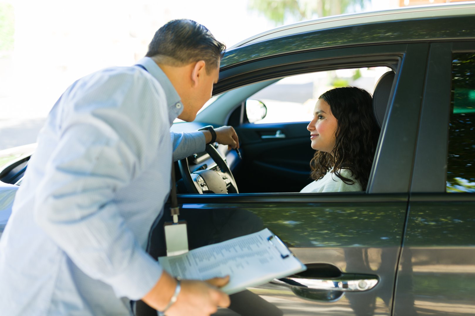 Latin driving instructor giving instructions to a teen girl during a driving test lesson while holding a clipboard