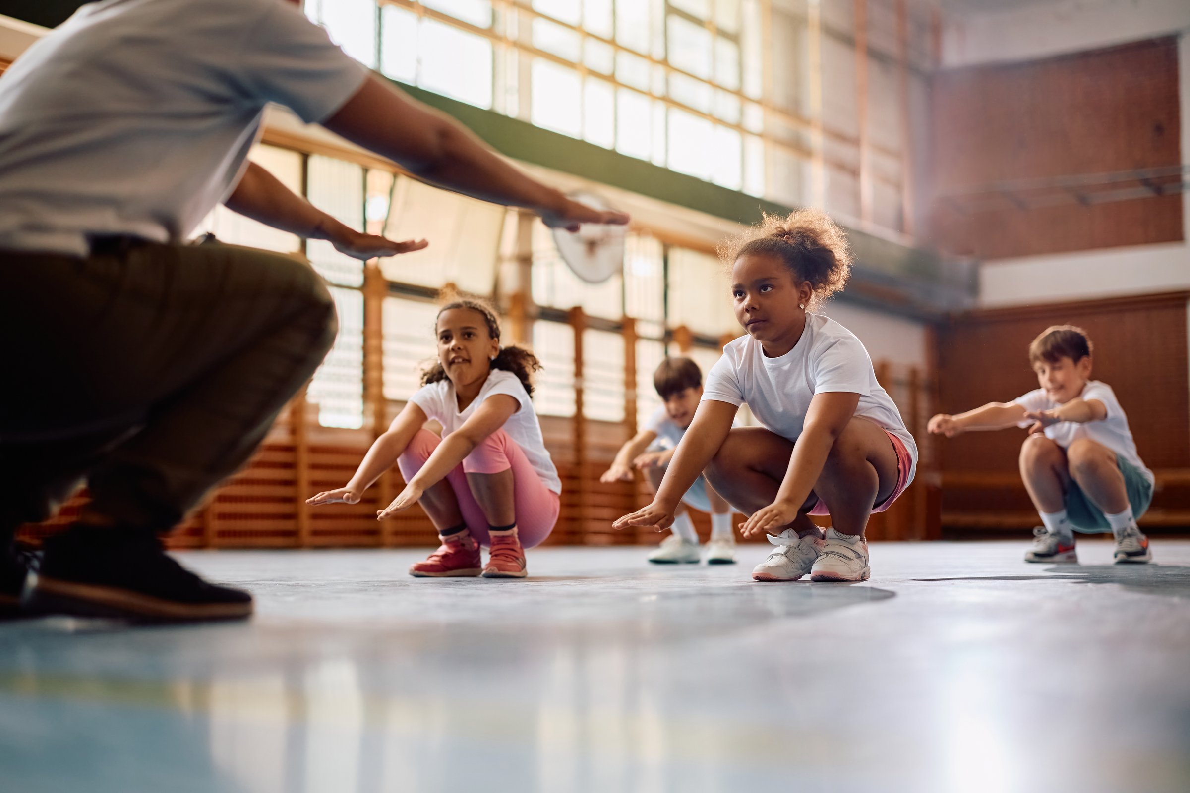 Group of elementary students exercising squats while having PE class with a coach at school gym. Focus in on African American girl.
