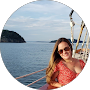 Woman with long hair wearing a red dress and sunglasses stands on a boat, with a scenic view of the sea and hills behind.