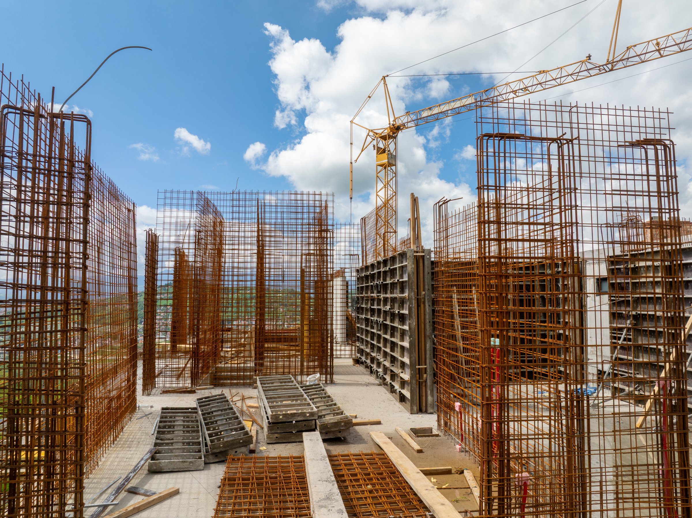 This construction site shows the skeletal framework of a building, with rebar, crane, and concrete forms under a cloudy sky, new structures being built.