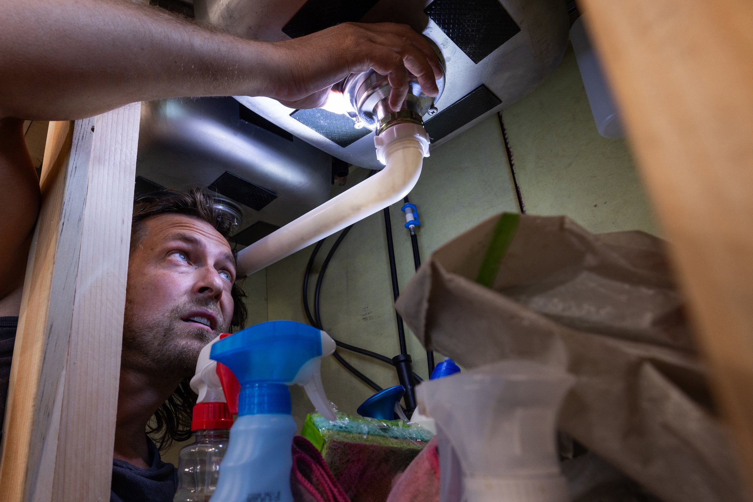 A man uses a flashlight to inspect or repair plumbing beneath a kitchen sink, surrounded by household cleaning supplies. This candid, real-life scene highlights home maintenance, DIY repairs, and responsible homeownership.