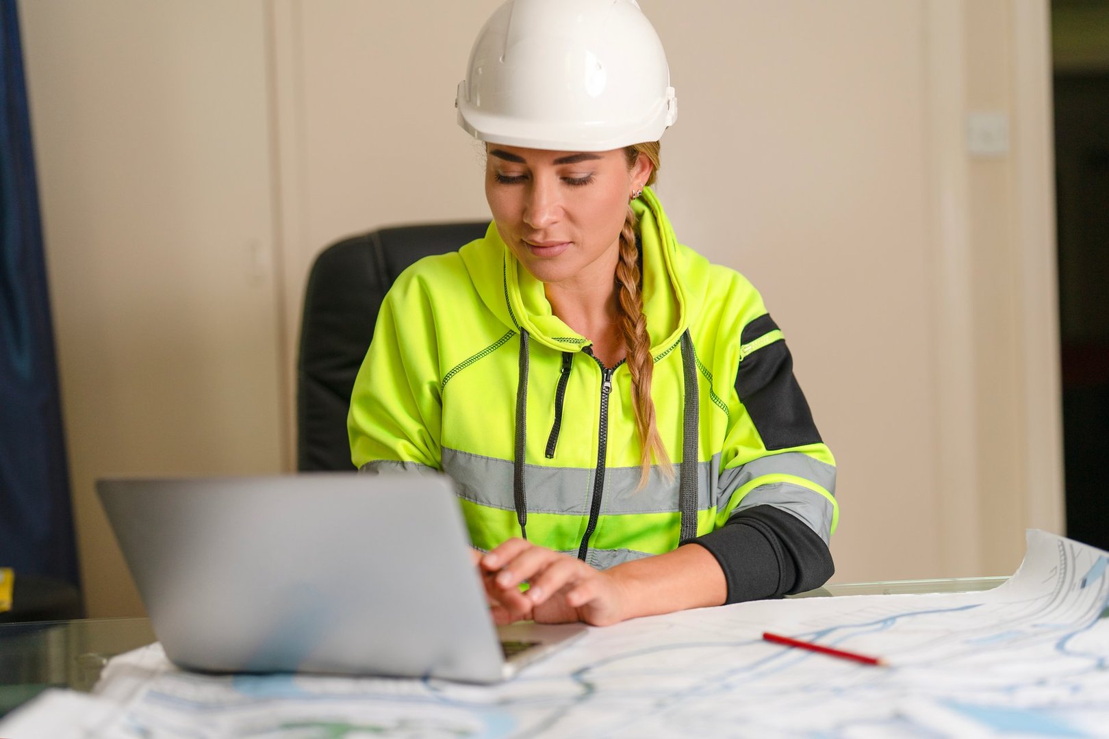 A construction engineer dressed in high-visibility clothing and a hard hat focuses on a laptop while reviewing detailed site plans in a well-lit office setting.