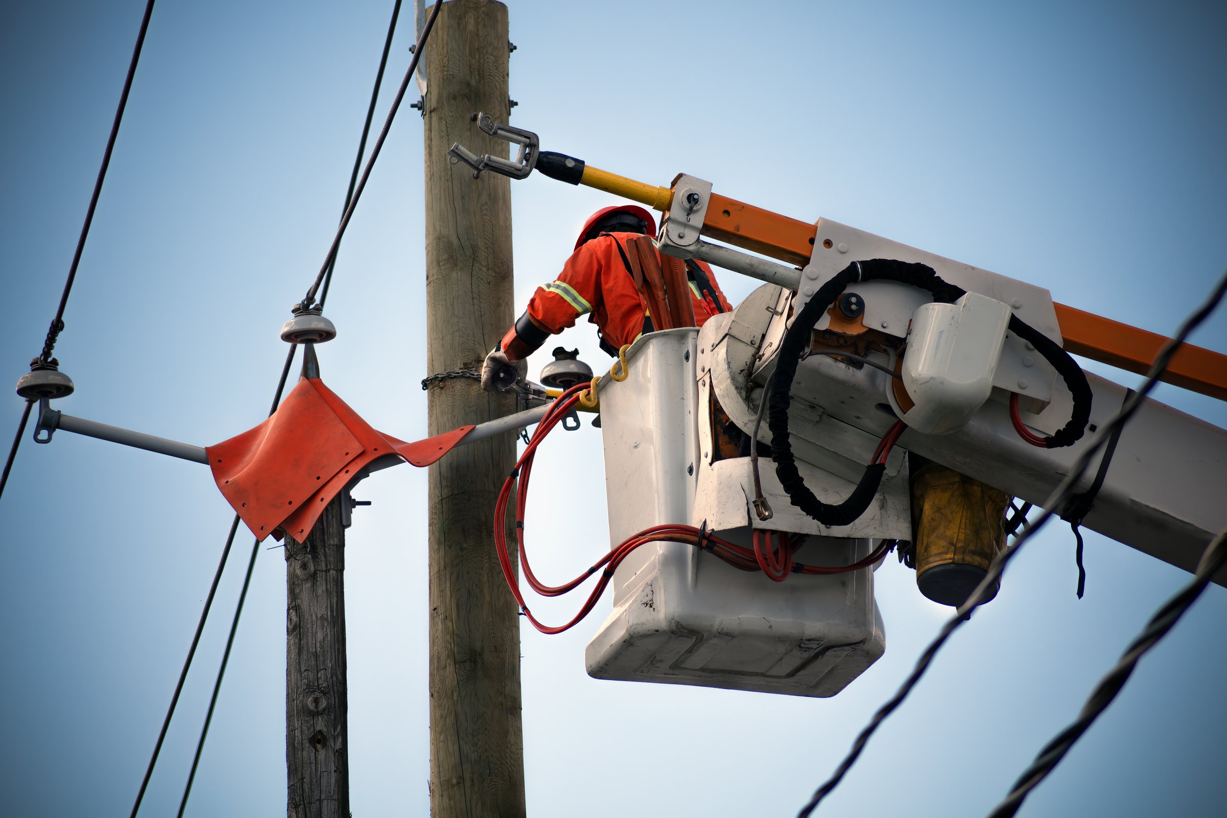 Power lineman performing maintenance on electrical lines from an elevated work platform. Essential for ensuring safe and reliable electrical service. Wearing orange high visibility vest and hat on blue sky fixing cables.