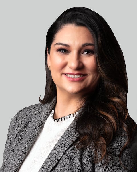 Smiling woman with long dark hair wearing a gray blazer and white top against a light gray background.