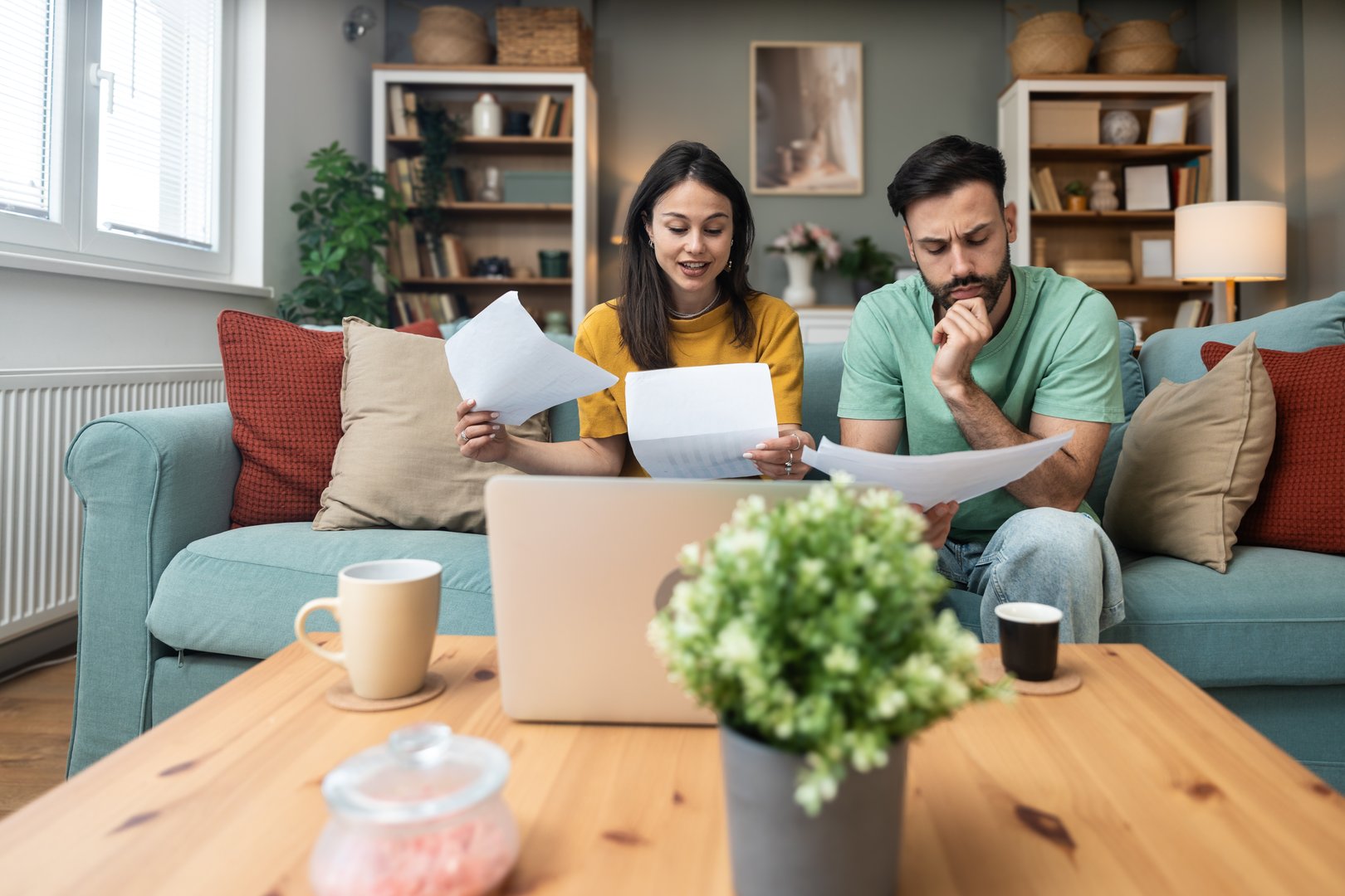 Worried young couple sitting on sofa at home, analyzing financial documents and statistics on laptop and paper, stressed about unexpected expenses, debt, or negative budget report results.