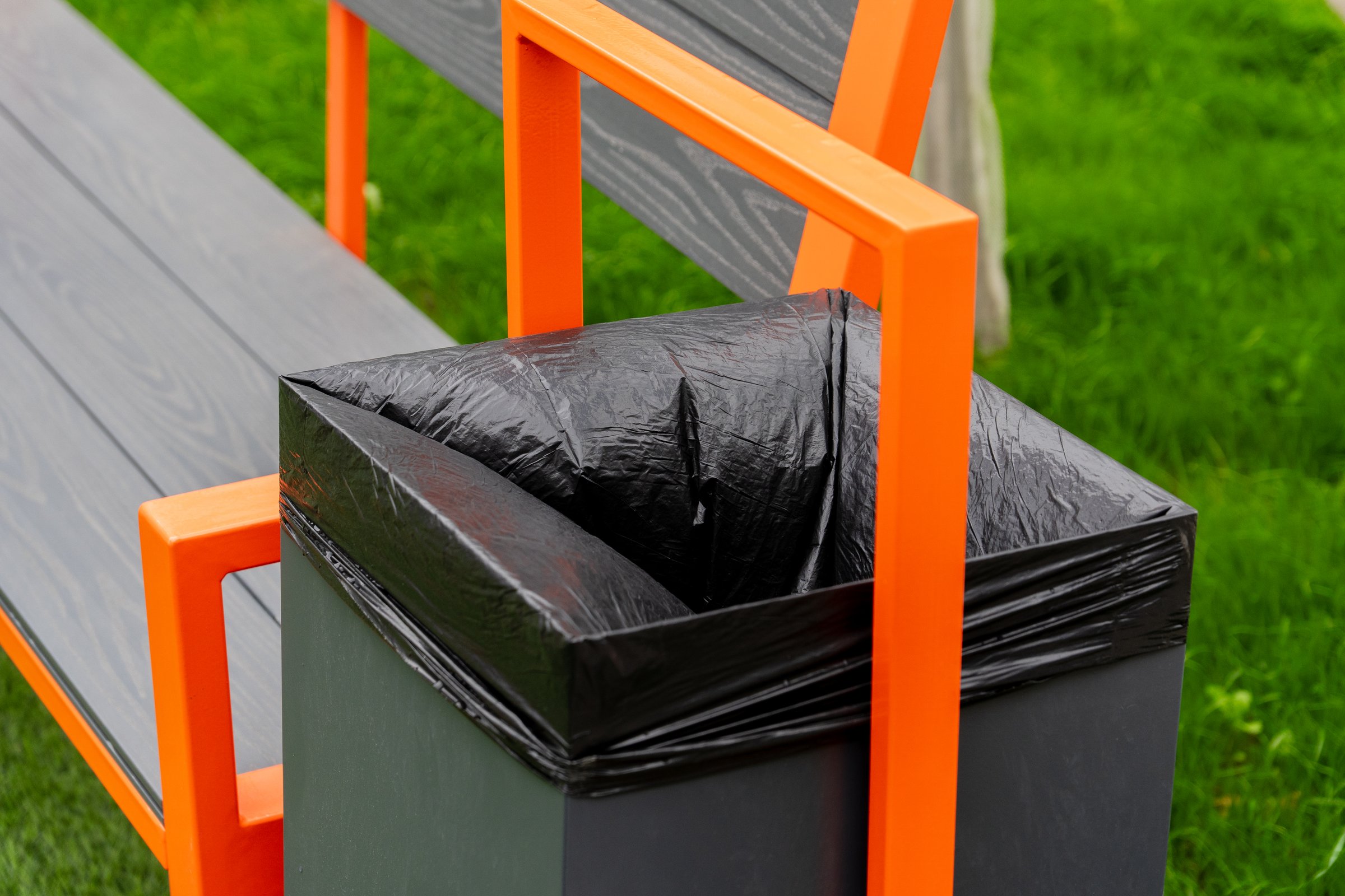 A modern bench featuring a built-in garbage container with a black liner sits on lush green grass, showcasing an aesthetic approach to waste management in public spaces.