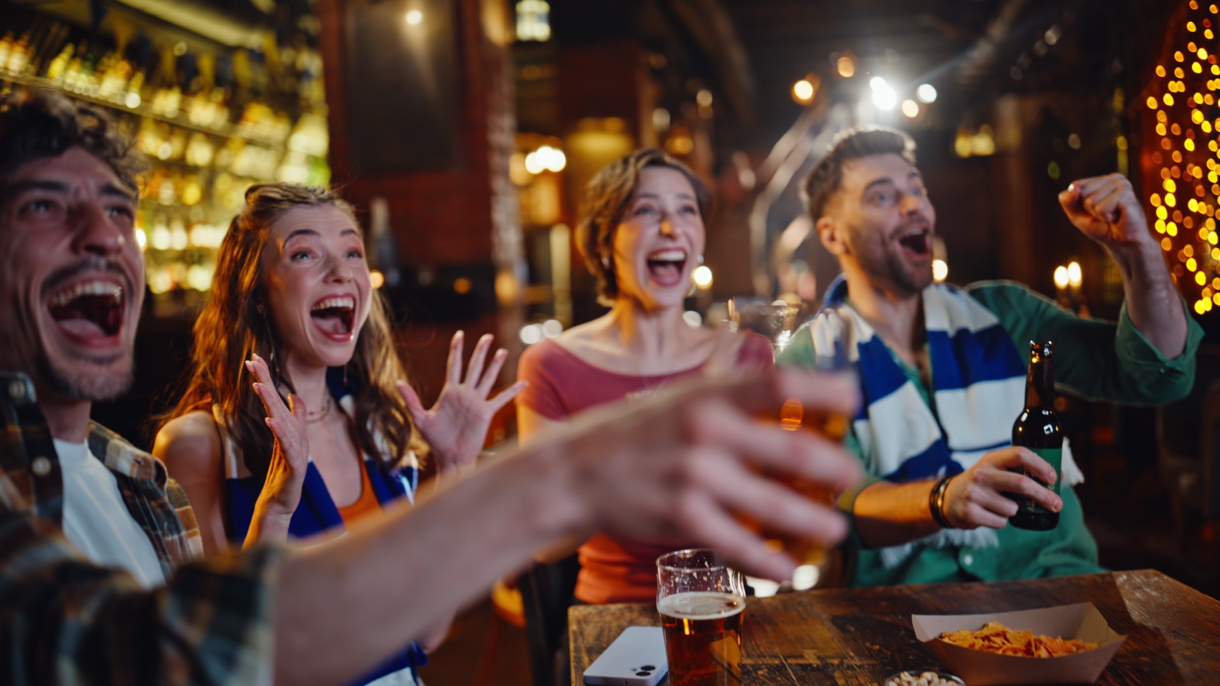 Excited students cheering sport game in modern pub drinking beer closeup. Happy group football fans watching competition on bar television. Overjoyed friends clinking glasses enjoying match at night.