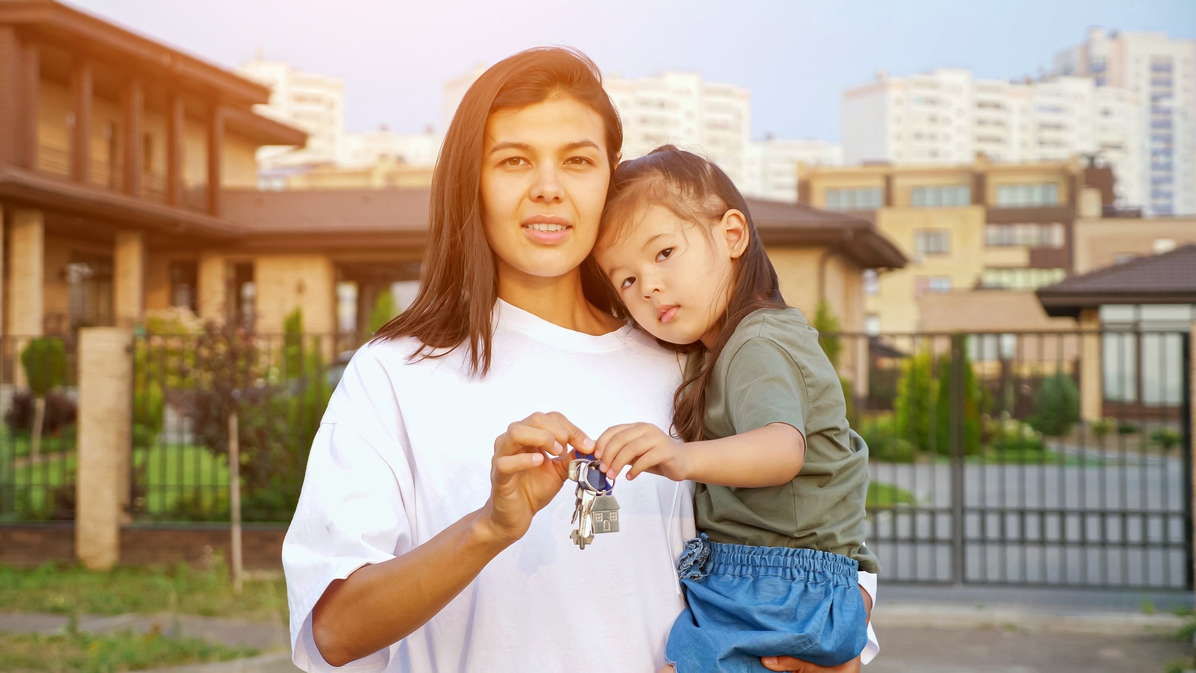 Cheerful woman shows keys of new apartment holding little Korean daughter in arms against detached house in city dwelling district closeup, sunlight
