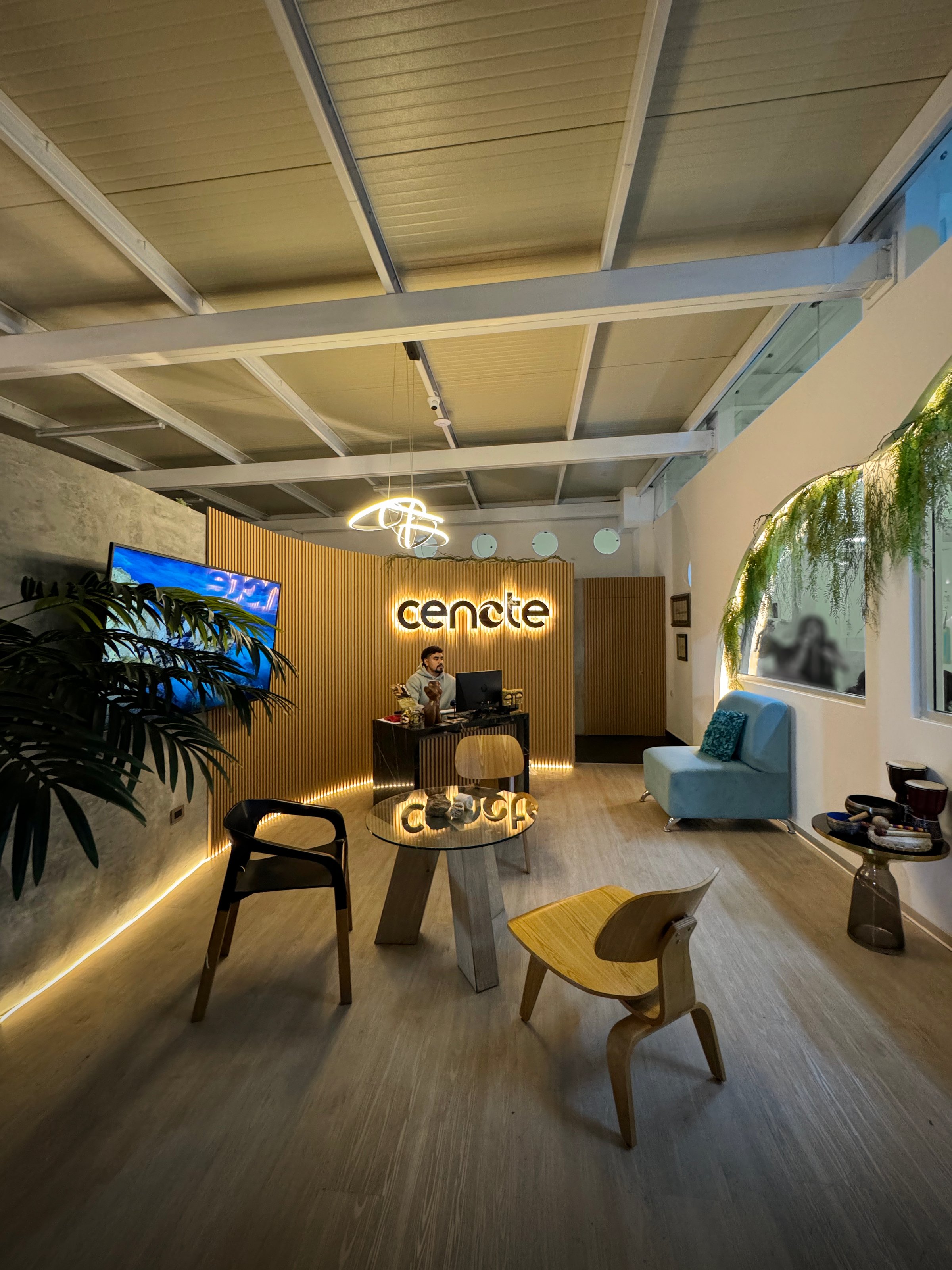 Modern office lobby with a reception desk, wooden decor, seating area, and ceiling lights. Person working at the desk.