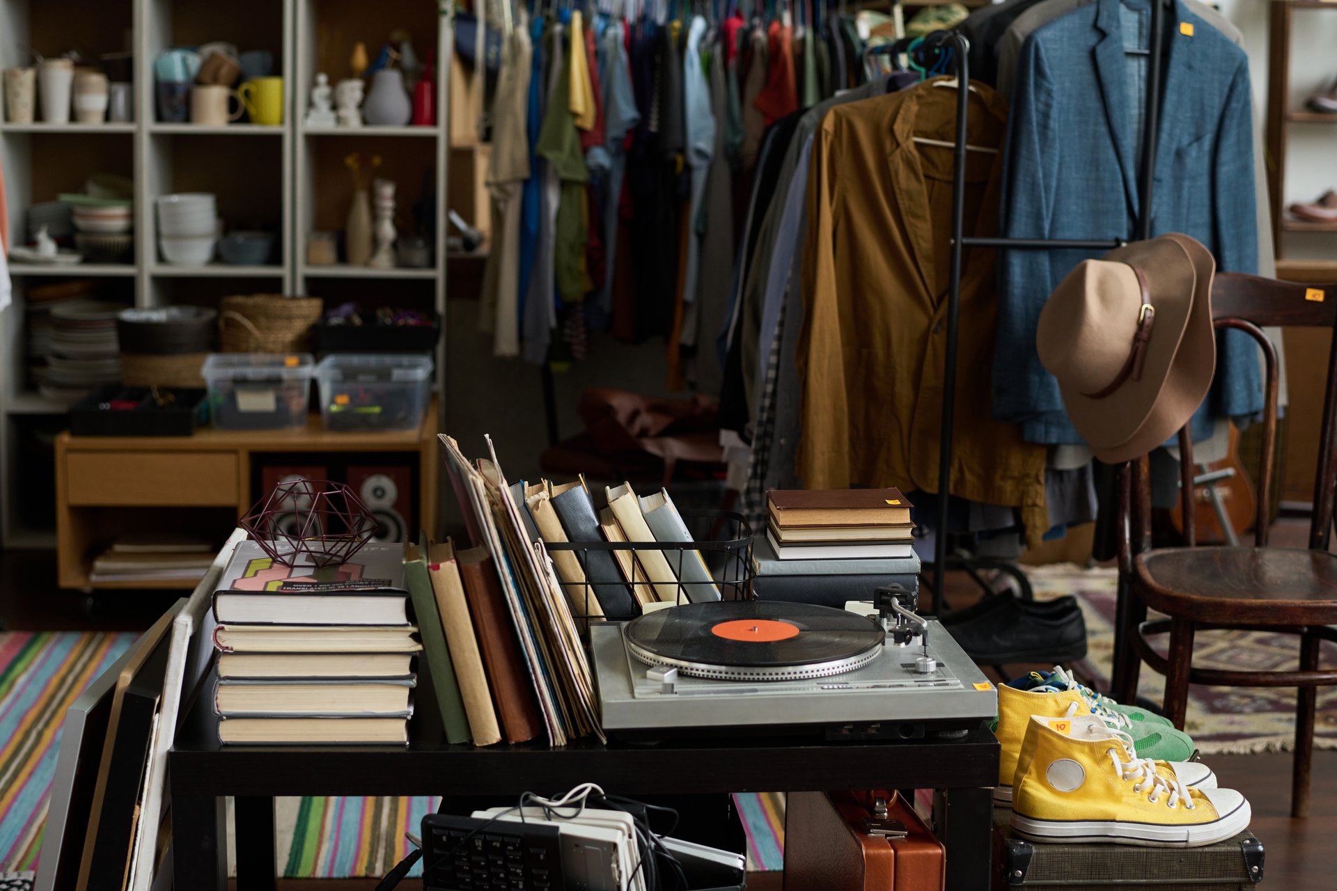 Several stacks of books, stereo record player on table standing by suitcases with sports shoes on top against racks with attire