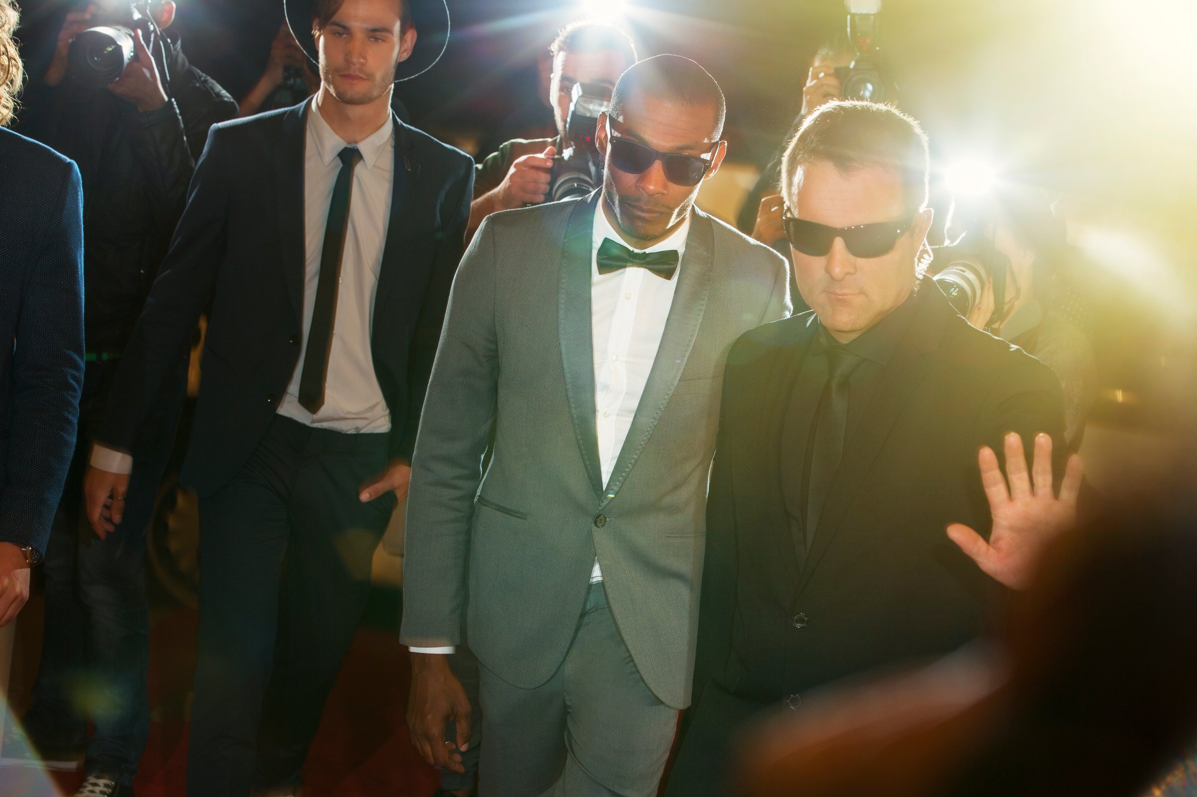 Two men in suits and sunglasses walk down a red carpet amid flashing lights. One holds up a hand to the cameras.