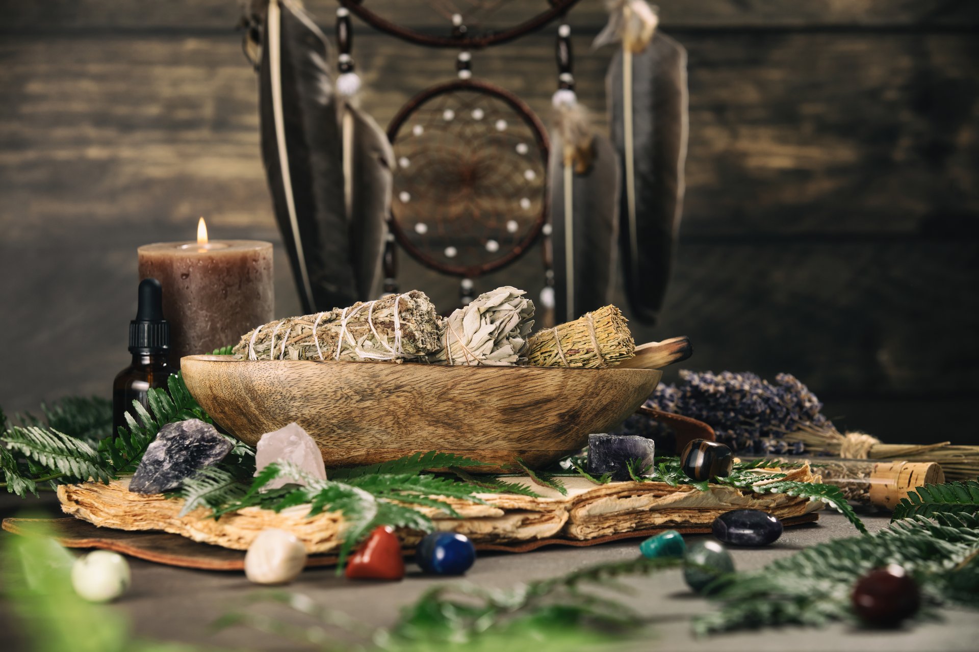 A serene arrangement of sage bundles, crystals, and ferns in a wooden bowl, with a lit candle and dreamcatcher adding to the spiritual ambiance.