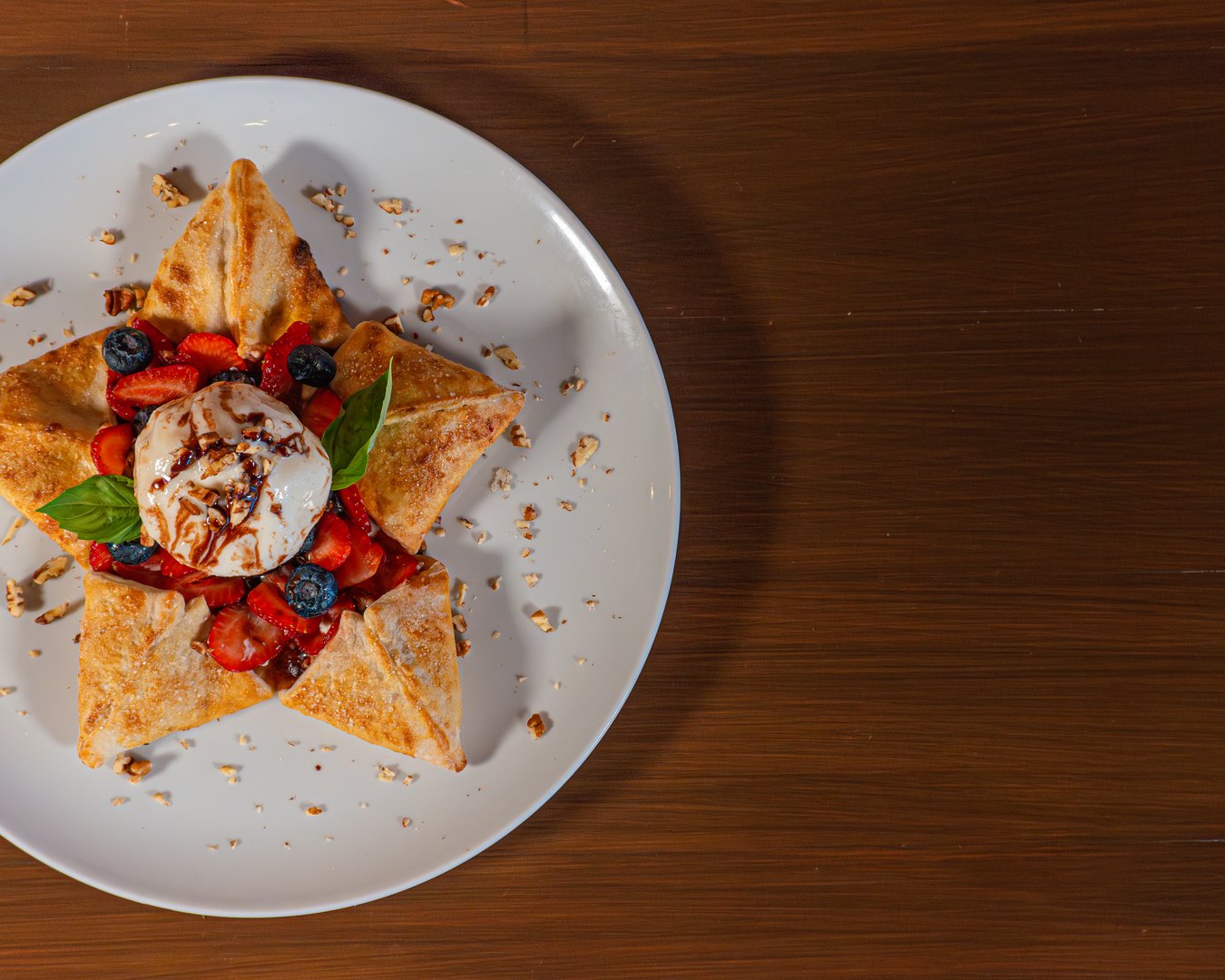 Elegant dessert of crepes topped with strawberries, blueberries, ice cream, and nuts, beautifully presented on a white plate