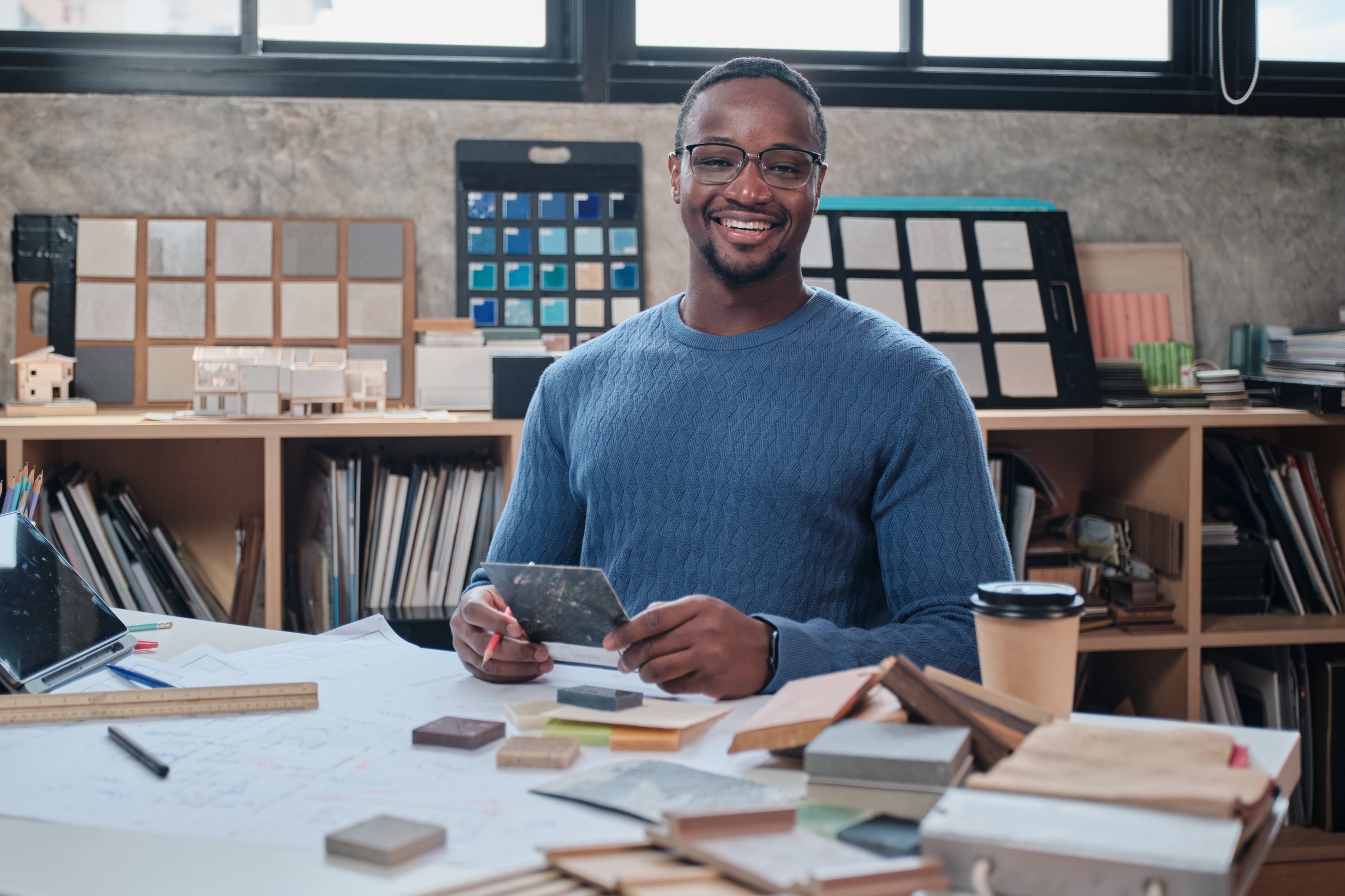 Portrait of Black male architect, happy design works on drawing blueprints and construction material on desk, smiles and looks at camera in company studio, creates ideas and inspiration concepts.