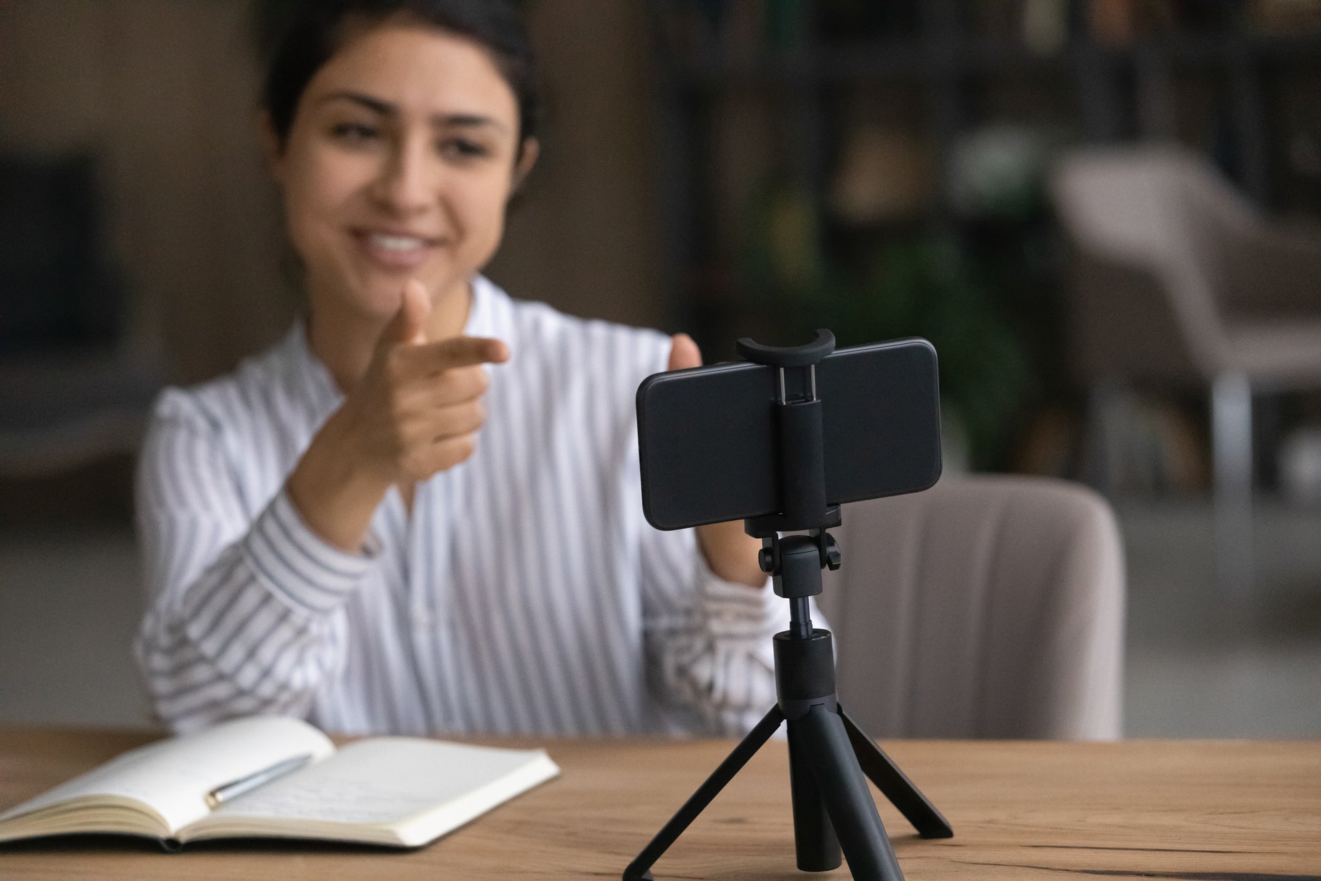 Video blogging. Active smiling young indian female shoot motivational training on cell webcam pointing fingers on gadget screen with enthusiasm. Focus on mobile phone on tripod at table used as camera