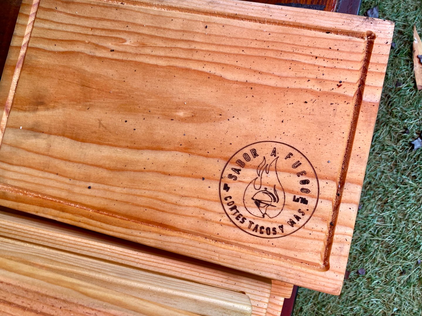 Wooden cutting board with a logo featuring a flame and text "Sabor a Fuego, Cotes Tacos" inside a circular stamp.