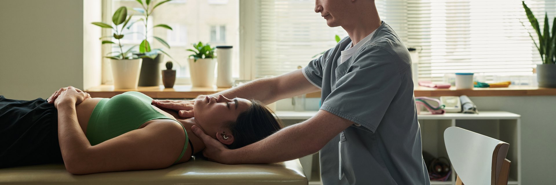 Health professional providing chiropractic treatment to female patient lying on massage table in well-lit clinic setting surrounded by potted plants and medical equipment