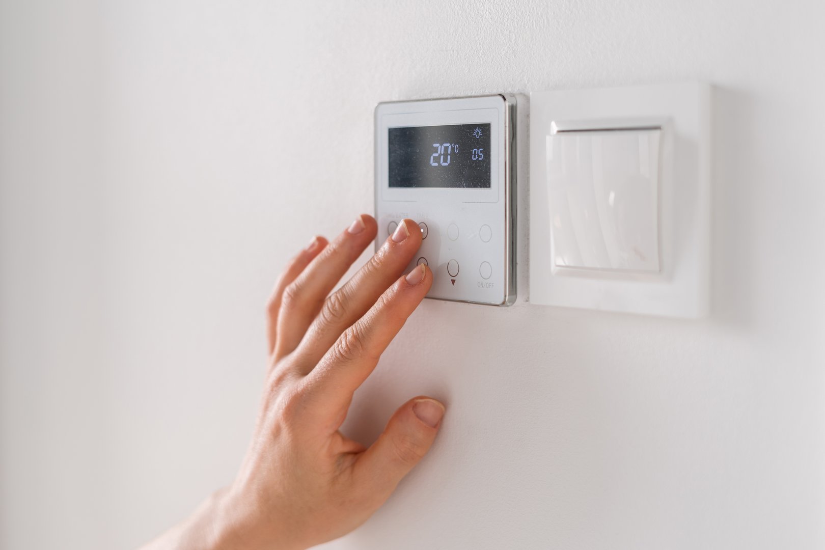 A hand adjusts the settings on a smart thermostat mounted on a white wall, optimizing temperature and energy use in a domestic living space for enhanced comfort and efficiency.