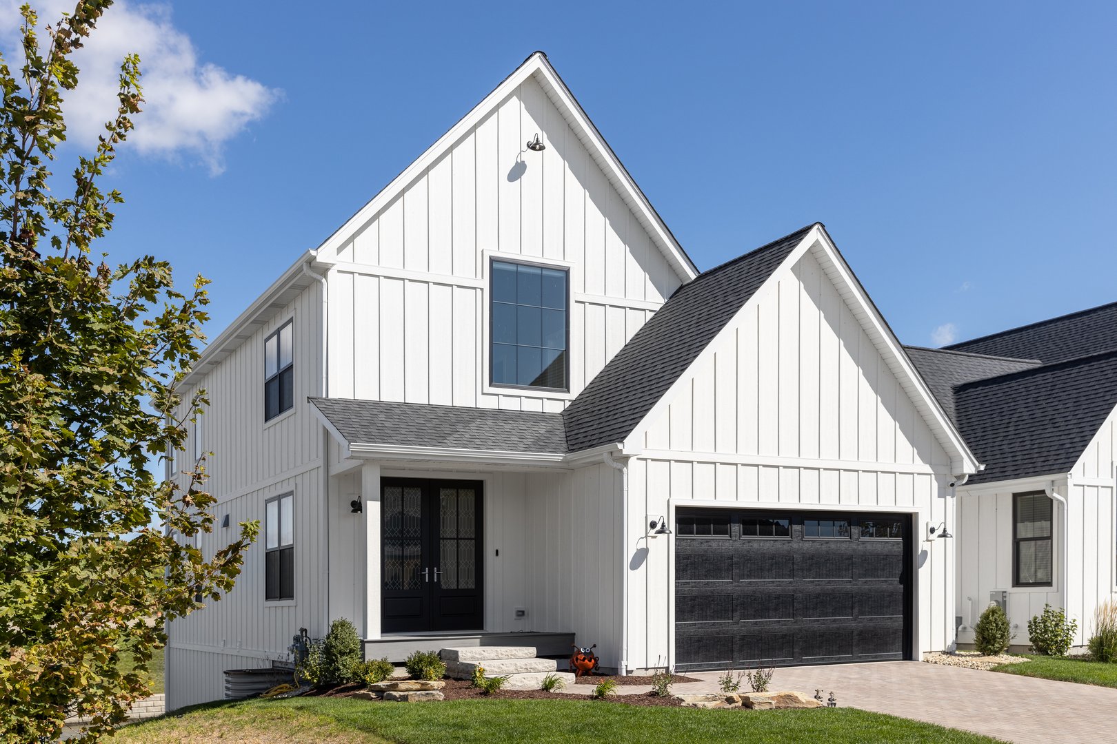 Elmhurst, IL, USA - September 26, 2022: A large, white modern farmhouse with vertical siding, a black two car garage, and lights mounted on the siding.