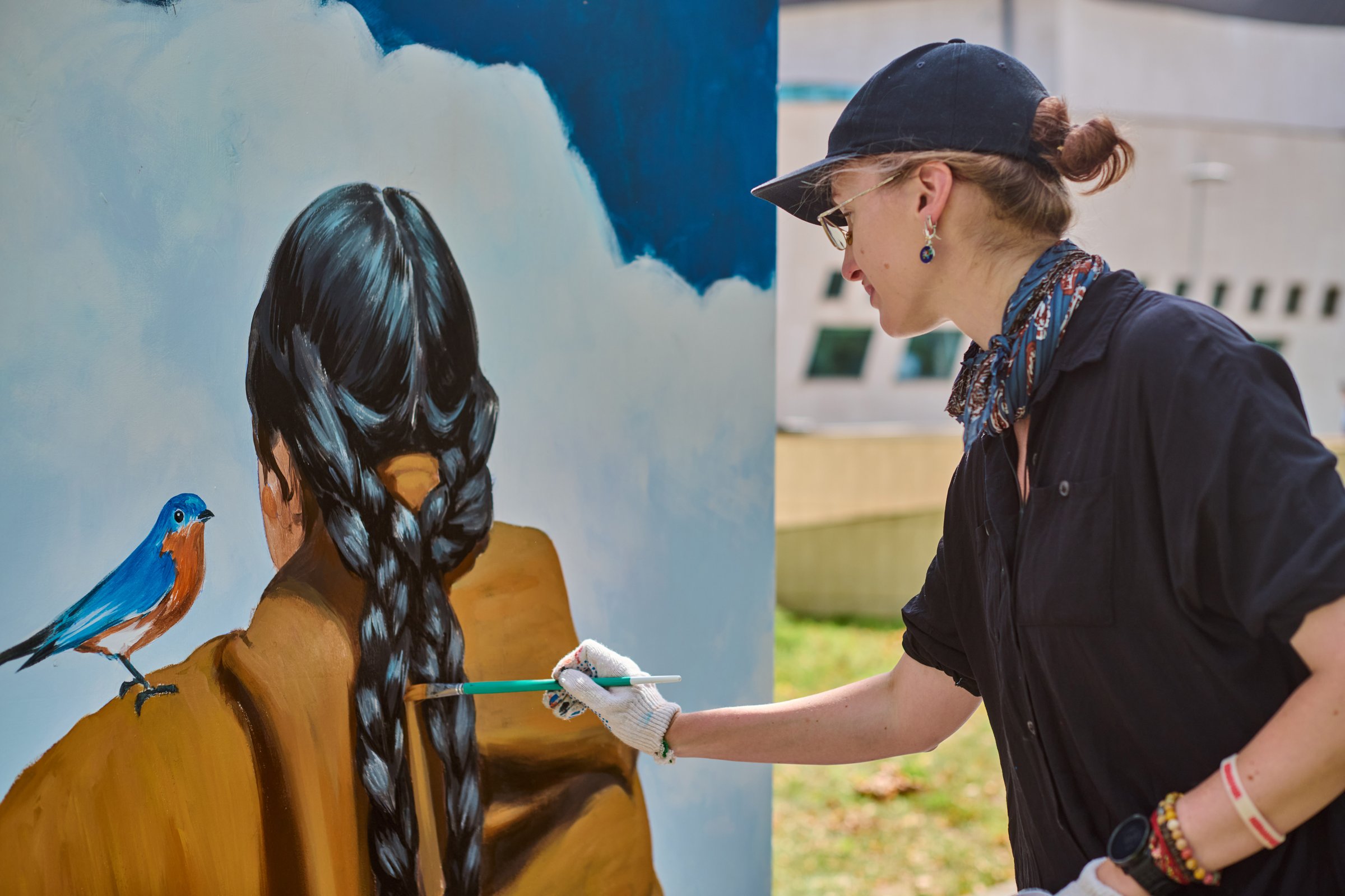 Young caucasian woman artist paints outdoor mural. Focus on braided hair figure, perched bird. Natural light, vibrant colors, dynamic scene emphasizes creativity