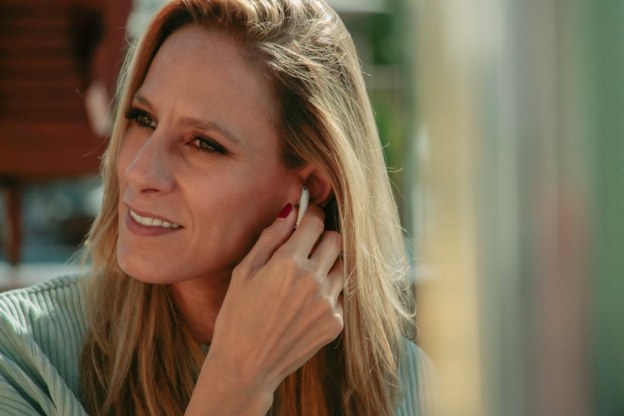 Woman smiling outdoors, adjusting an earbud with her left hand, wearing a light green top.