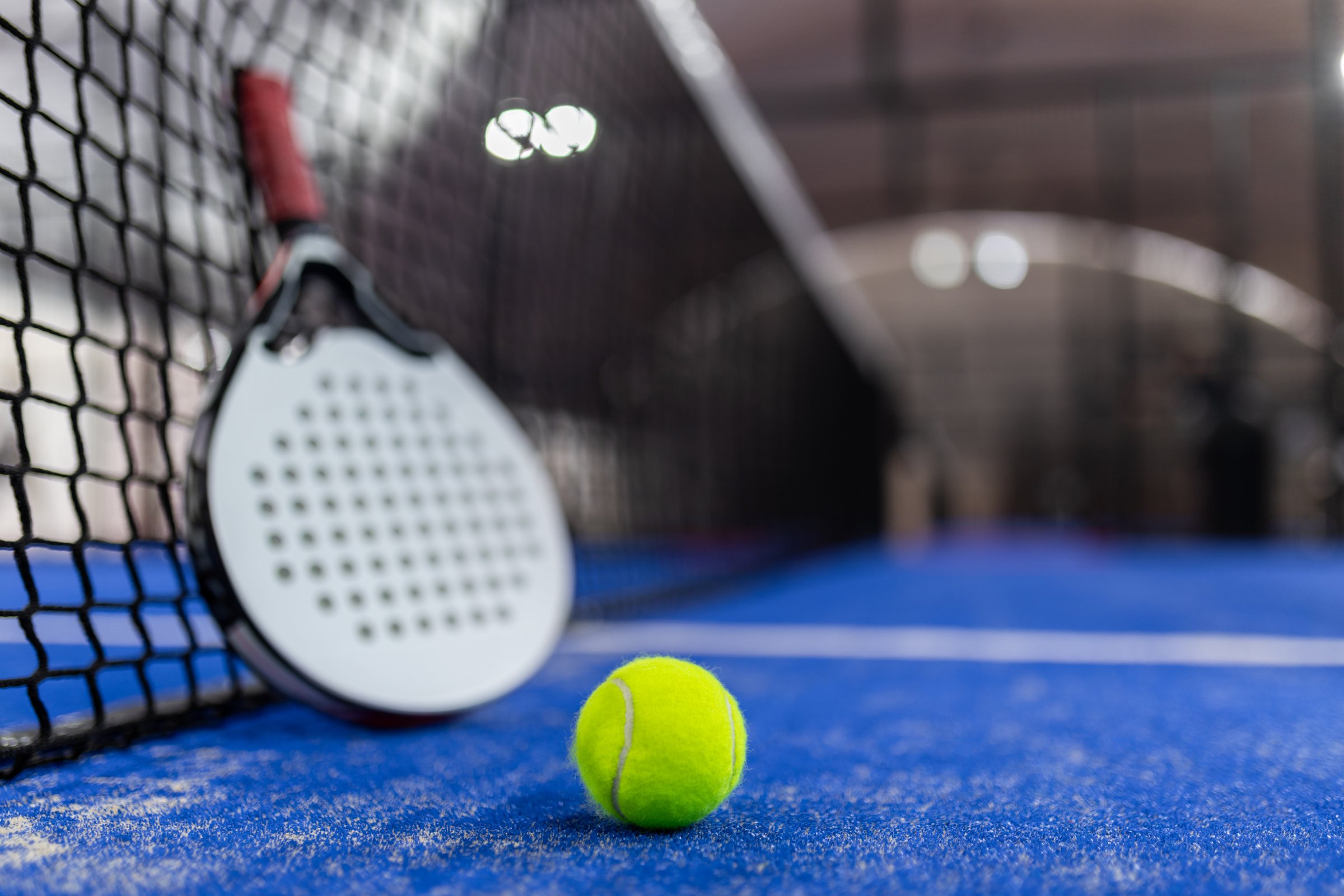 A padel racket lies against the net alongside a vibrant green ball, set on a blue surface of an indoor padel court, capturing the essence of an active sport.