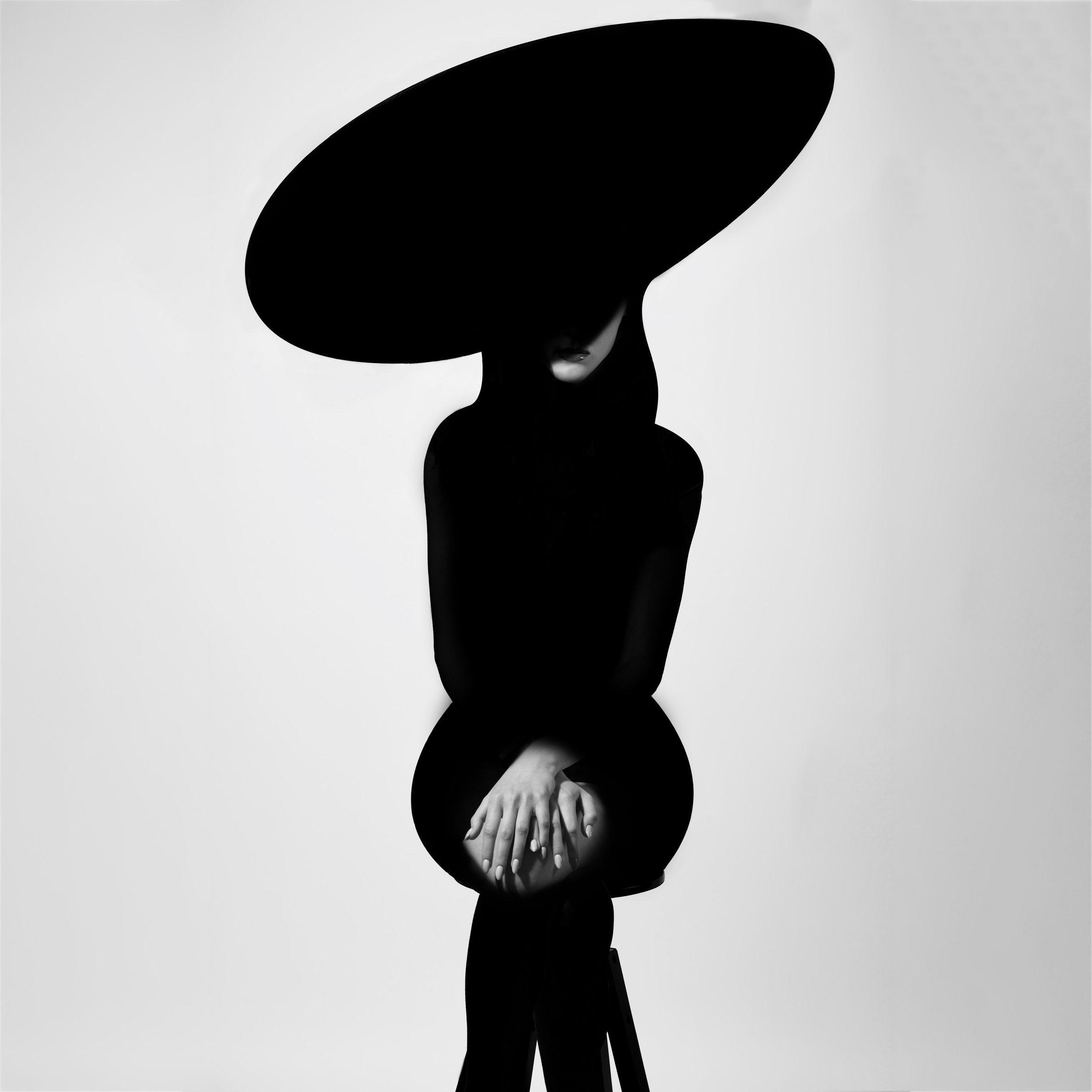 Fashion concept. Abstract woman black body silhouette with big hat and black clothes. Part of face and hands illuminated with light. Sexy model body shape. Sitting on chair in grey studio background